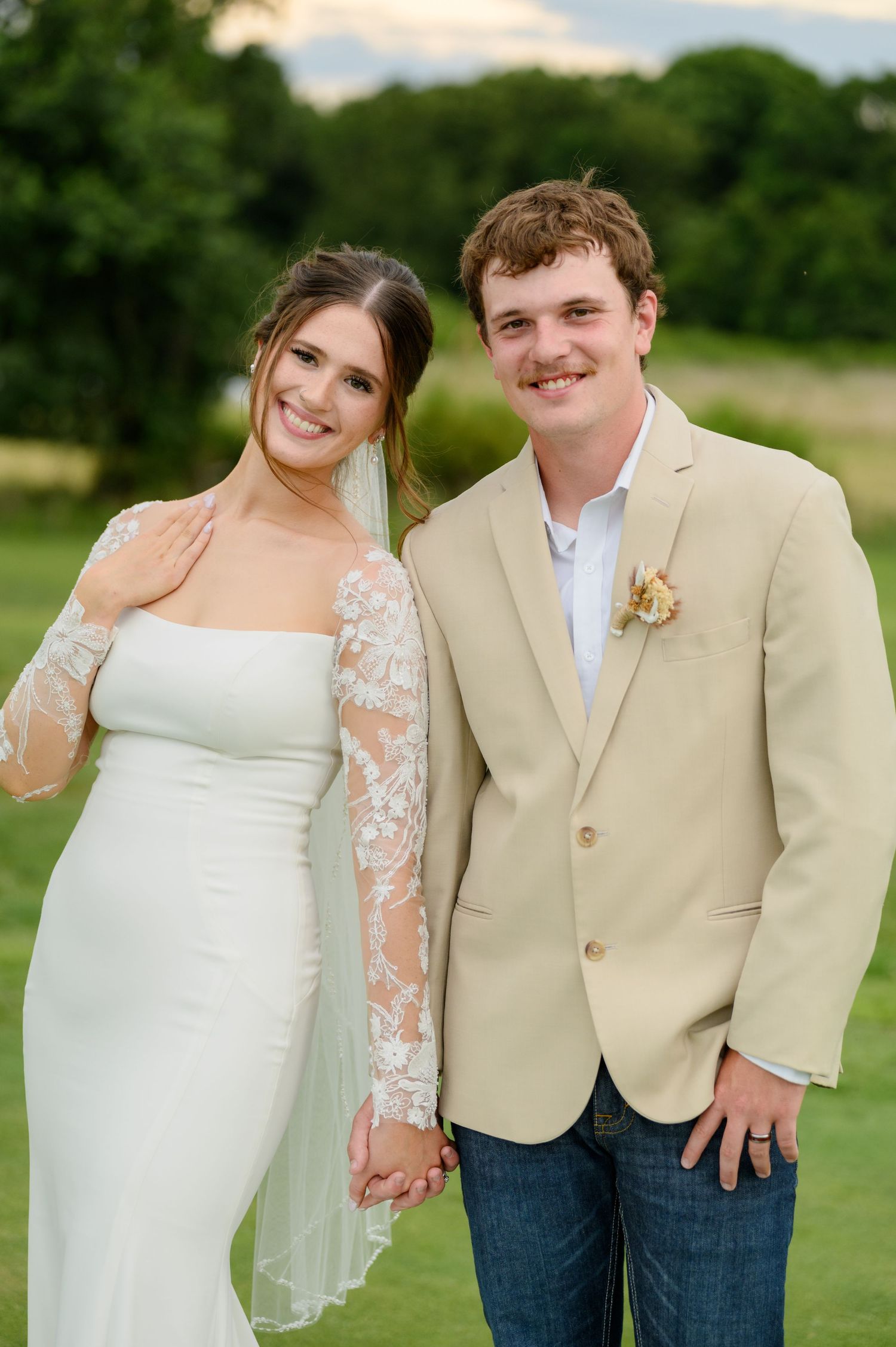 Bride in lace sleeve gown and groom in tan blazer holding hands smiling at outdoor wedding.