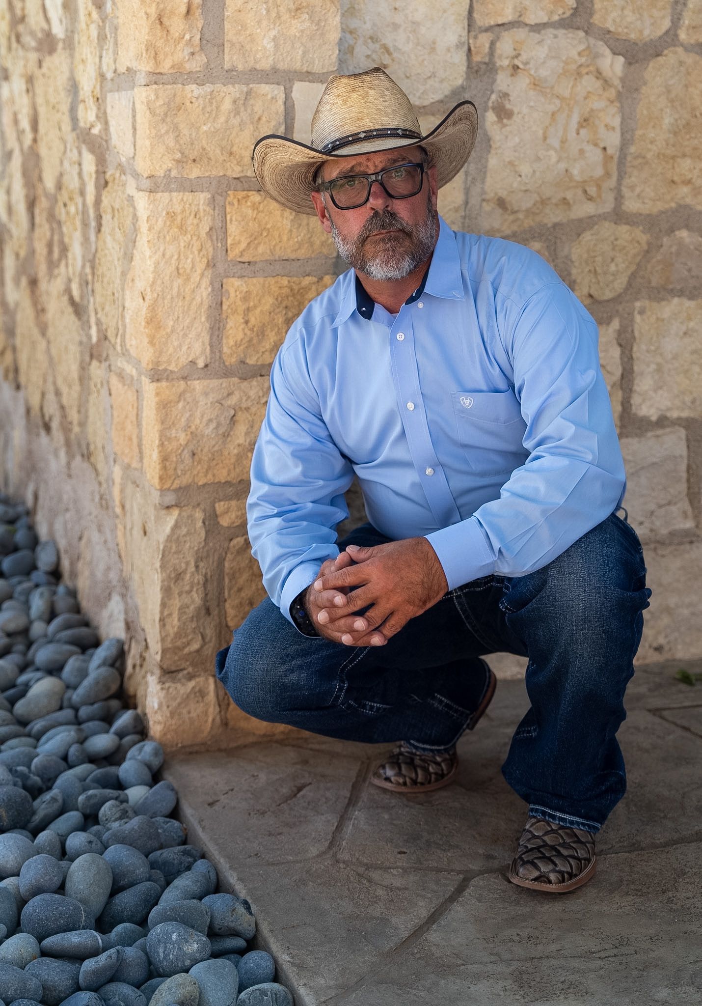 Man in cowboy hat, glasses, blue shirt, and jeans crouching by stone wall with river rocks.