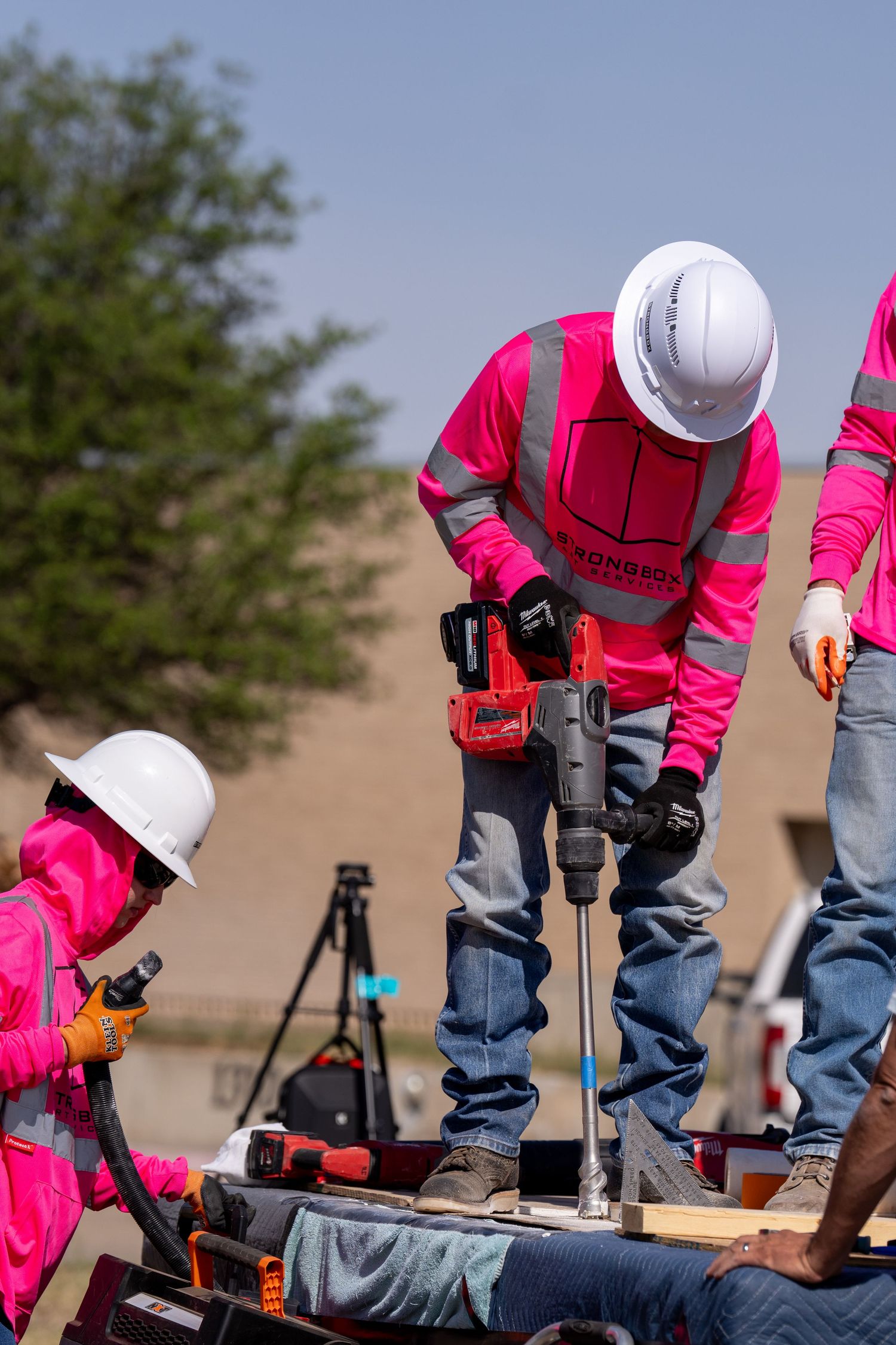 Construction workers in pink safety vests and white hard hats using a power drill on a job site outdoors.