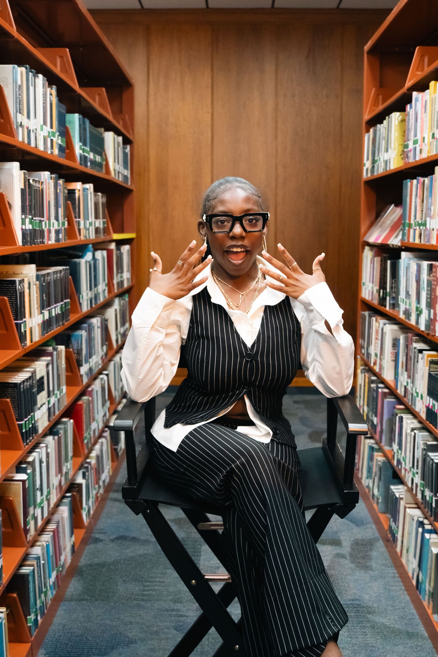 A woman in striped vest and glasses sits between library shelves with hands raised in a surprised expression.