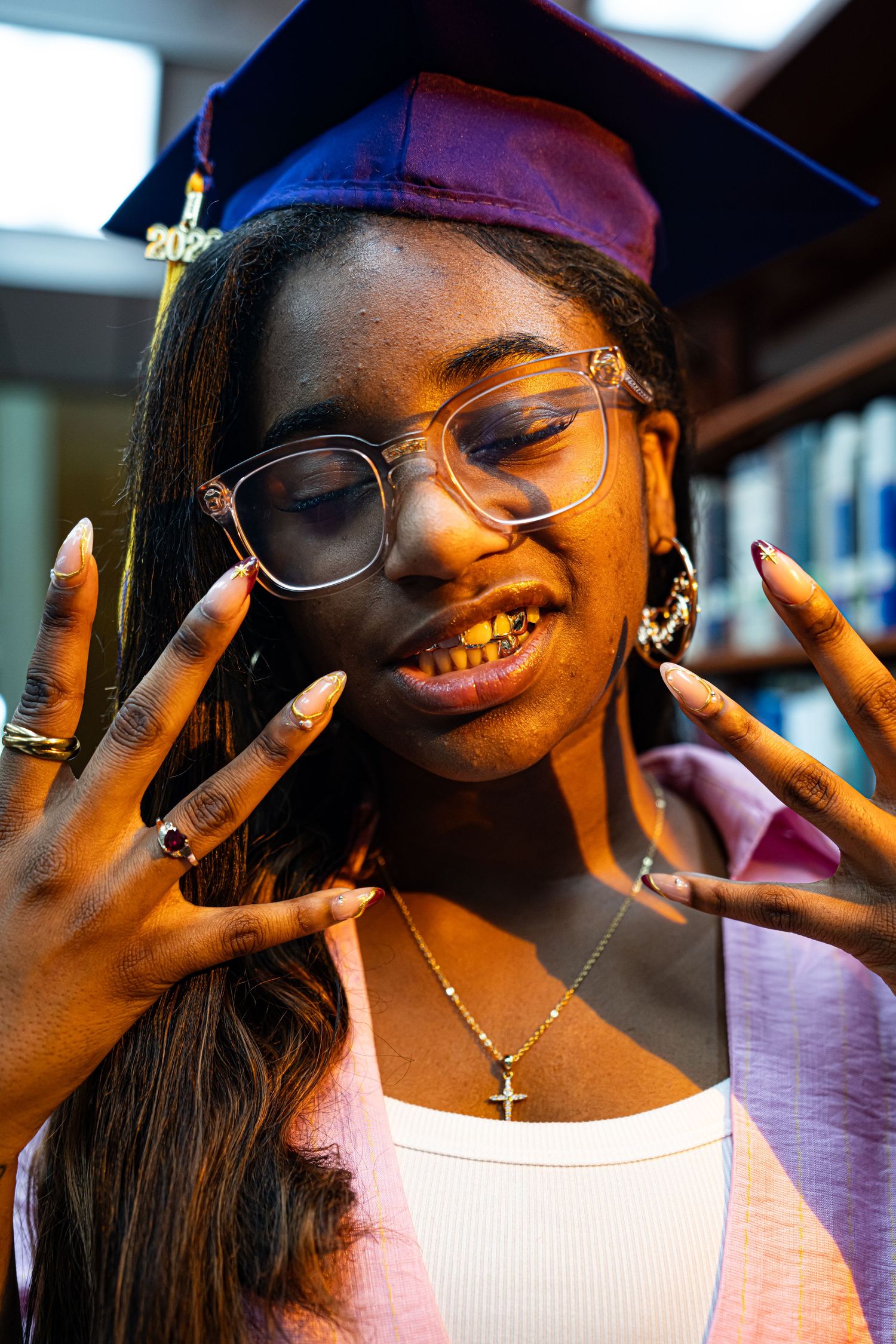 Young woman in graduation cap and glasses showing long nails, wearing gold grillz and cross necklace.