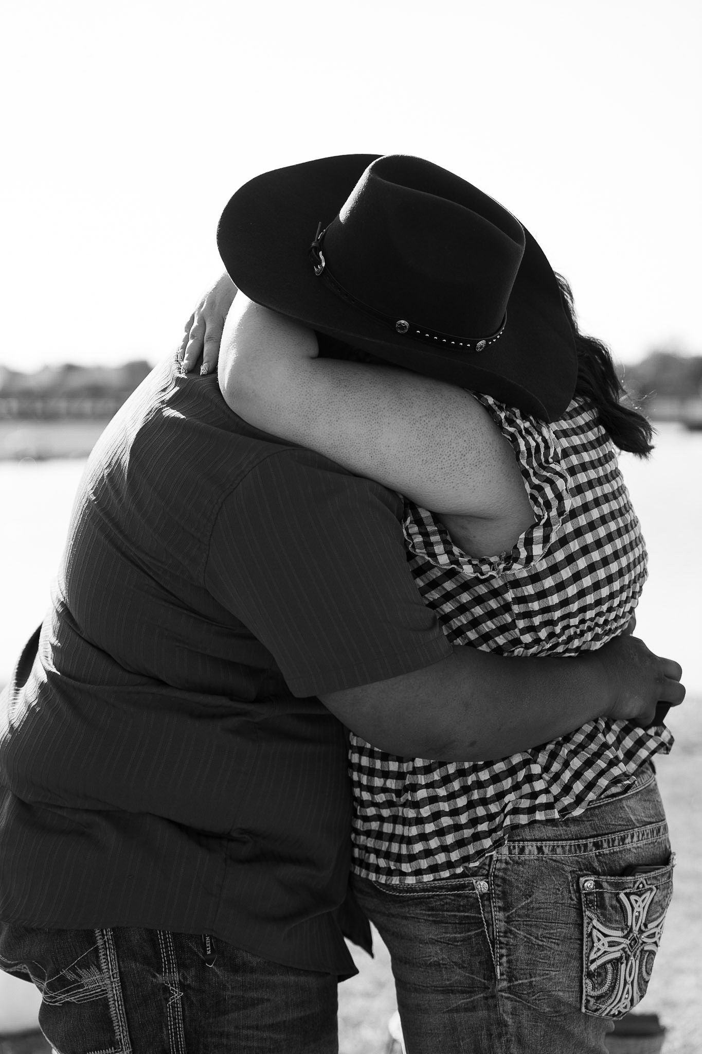 Couple shares emotional embrace in black and white photo near waterfront, one wearing cowboy hat.