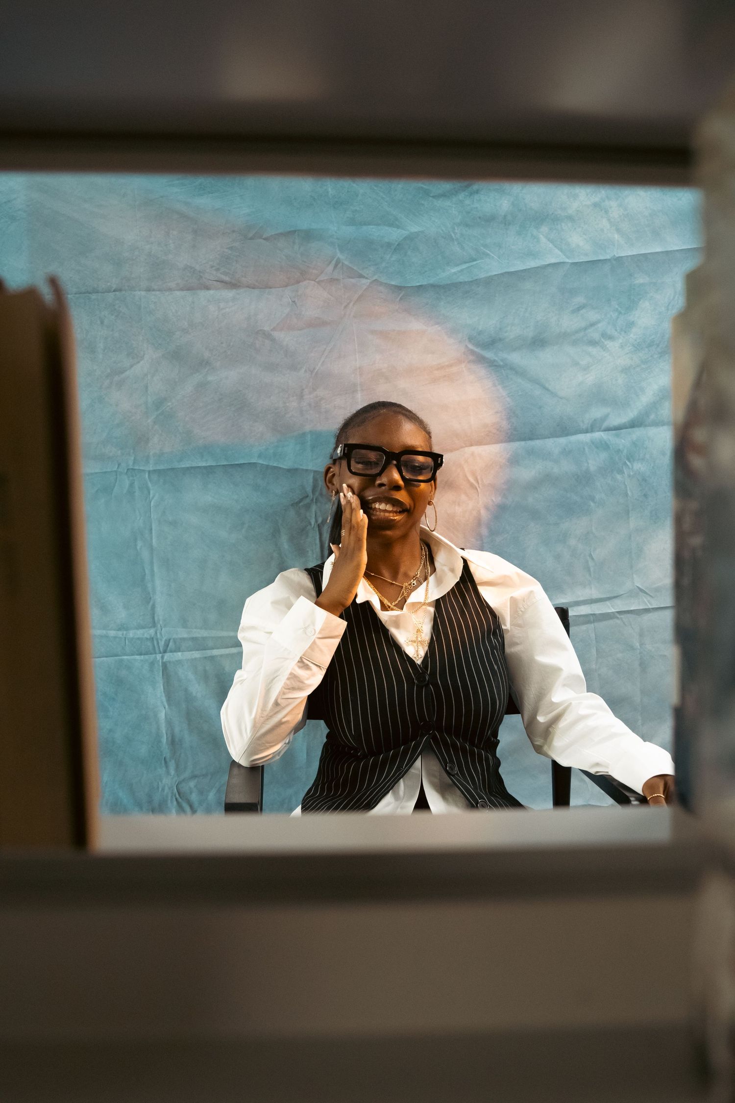 Black woman in pinstripe vest talks on phone seated in director chair against blue backdrop.