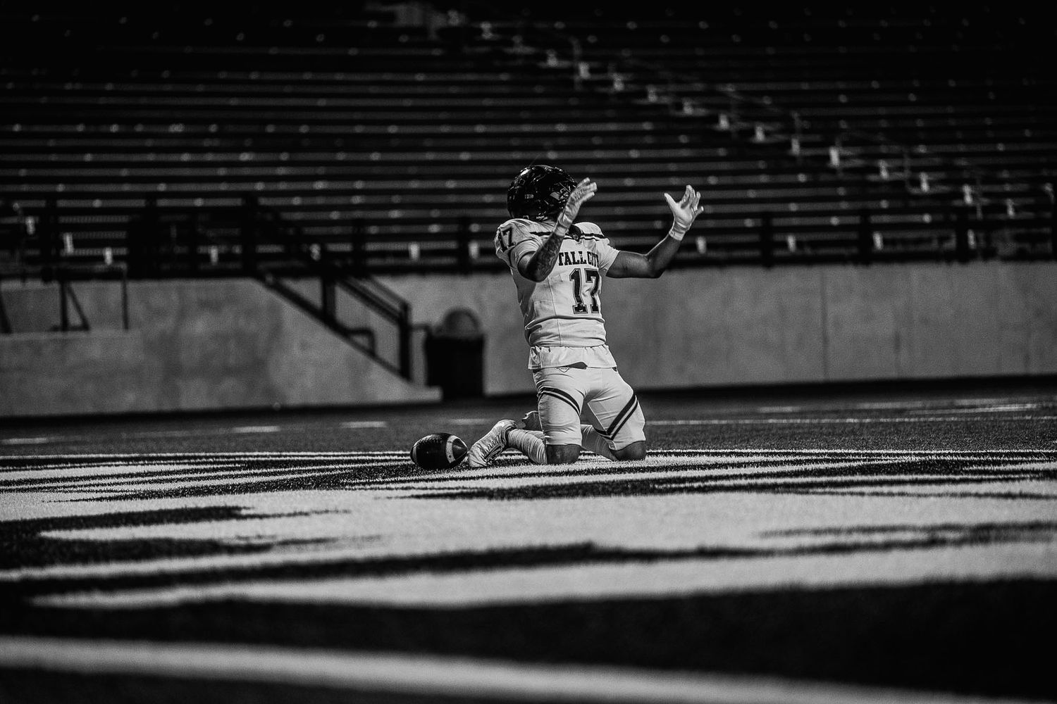 Football player #17 kneels on field in black and white photo inside empty stadium.