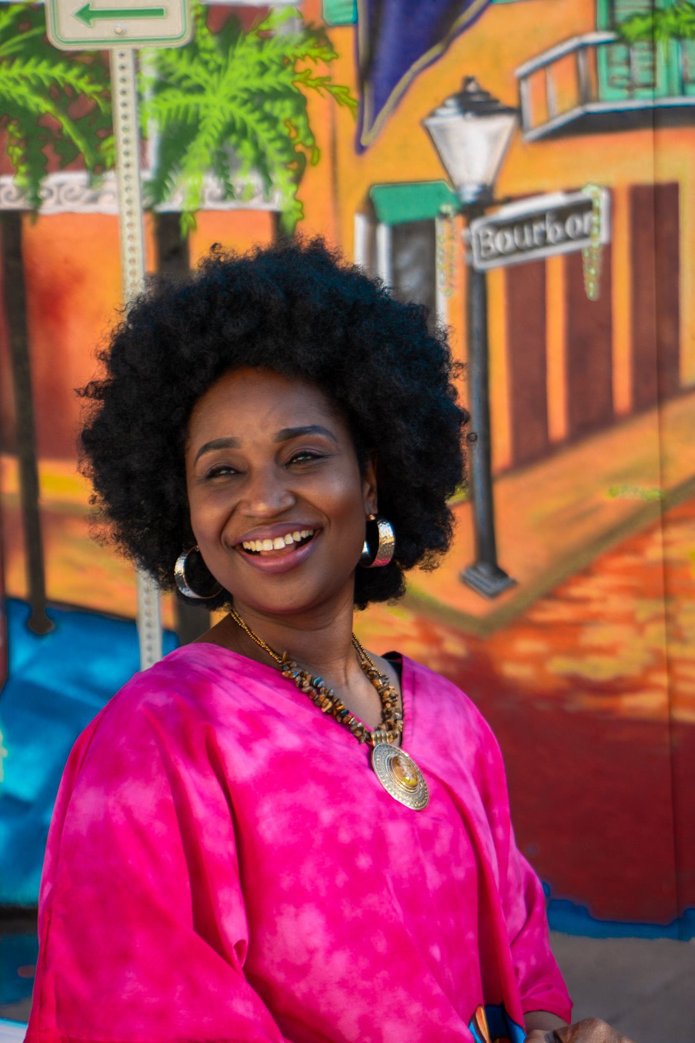 Smiling woman with afro in pink top poses before colorful Bourbon Street mural.