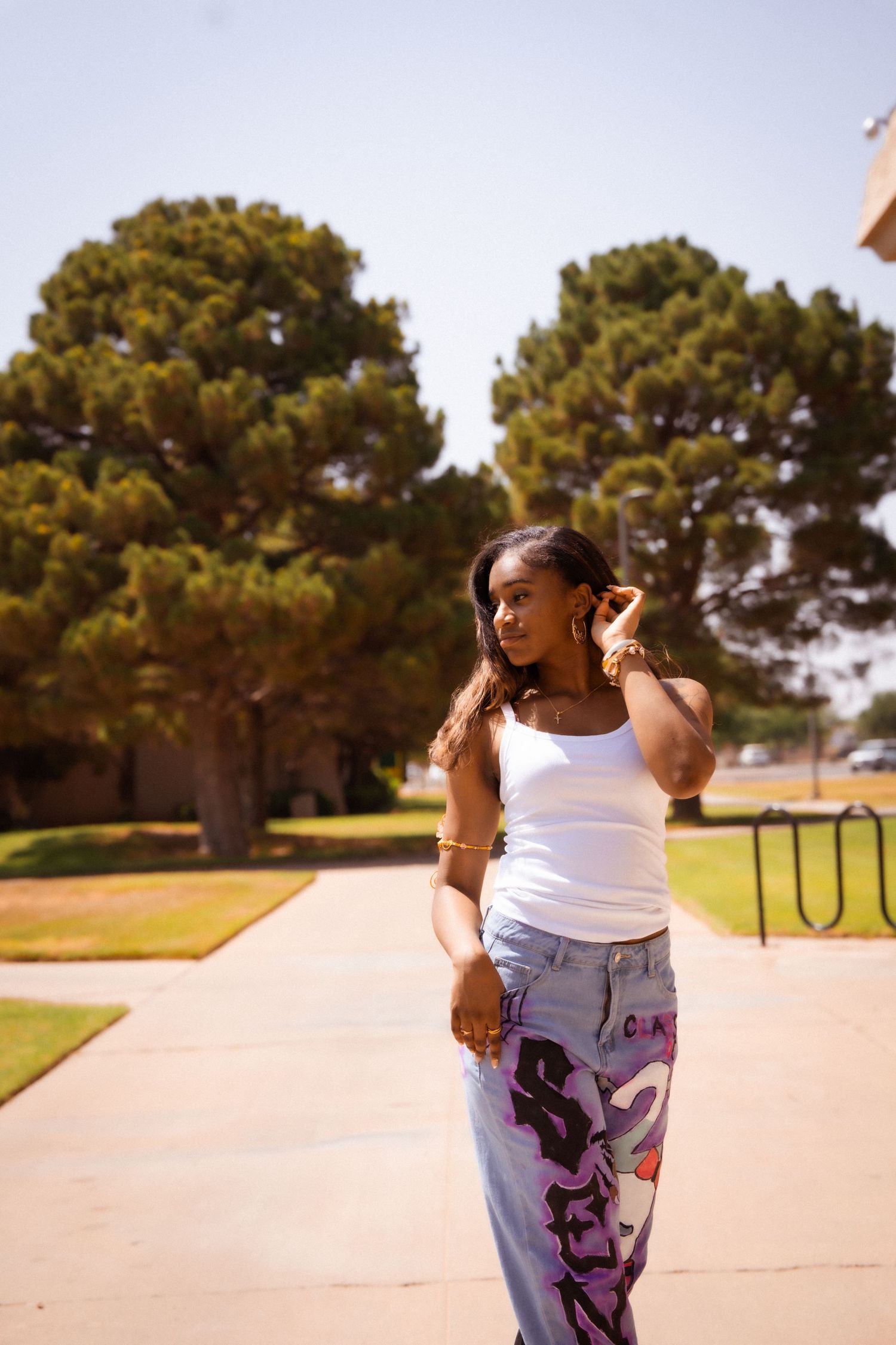 Young Black woman in white tank top and graphic jeans walking outdoors near trees on sunny day.