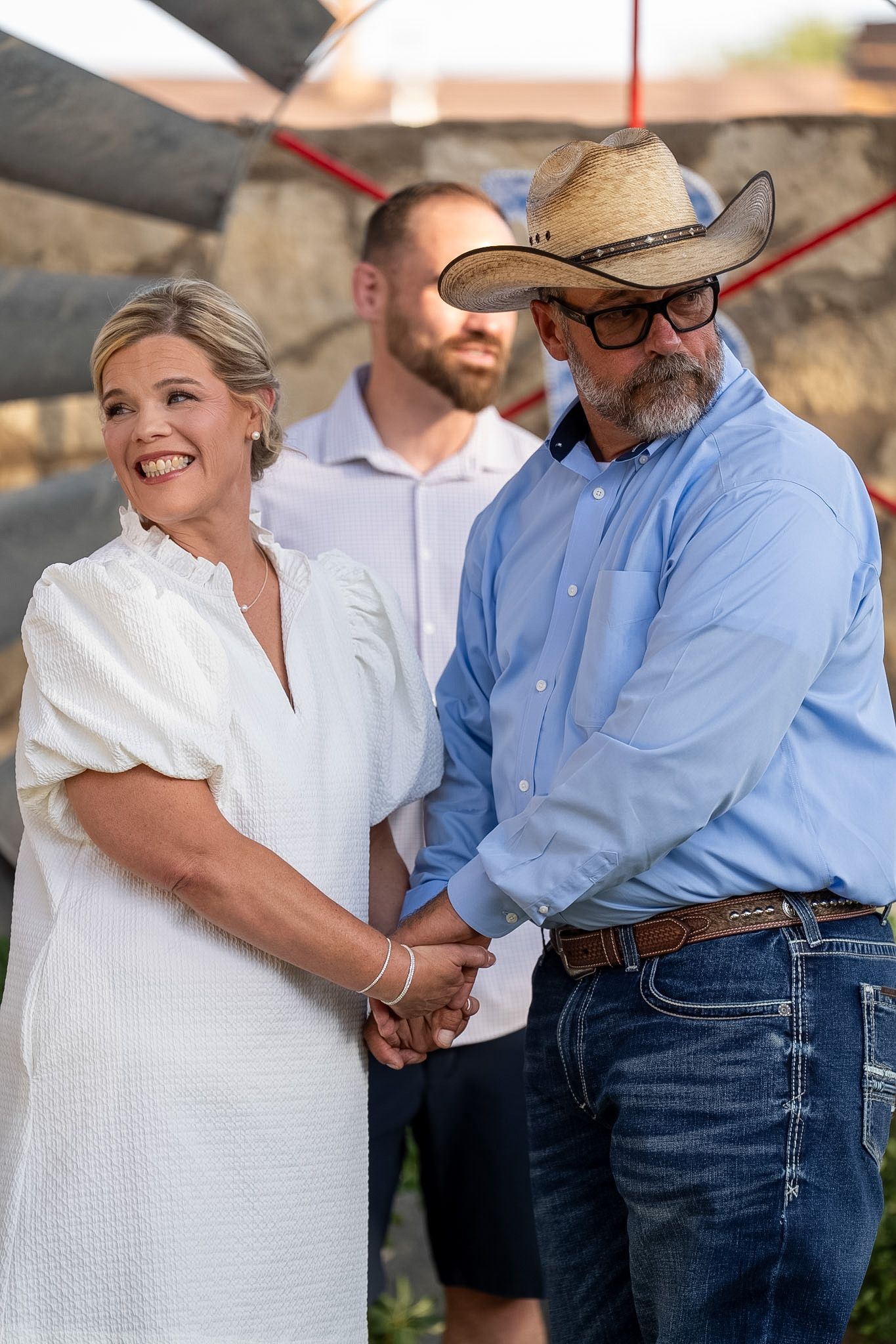Bride in white dress and groom in cowboy hat holding hands at outdoor wedding ceremony.