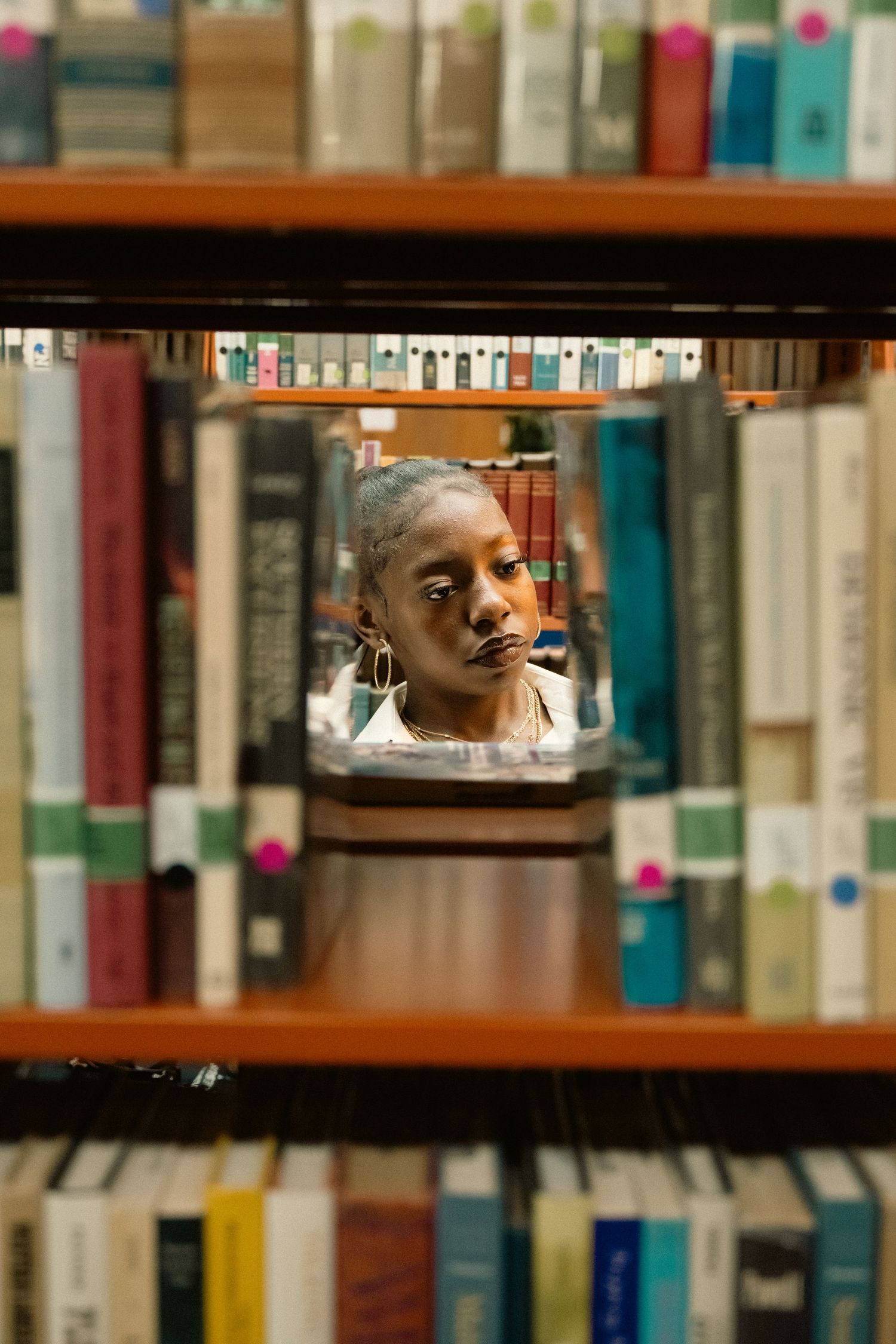 Woman with gray hair viewed through library bookshelf gap, surrounded by colorful books in a library.