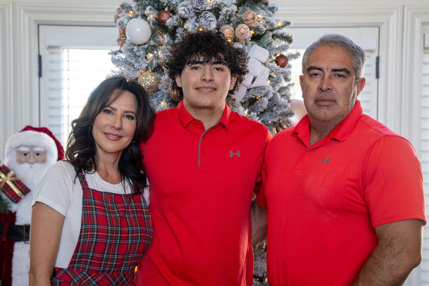 Family of three posing together in front of a decorated Christmas tree, wearing red outfits for a holiday photo.