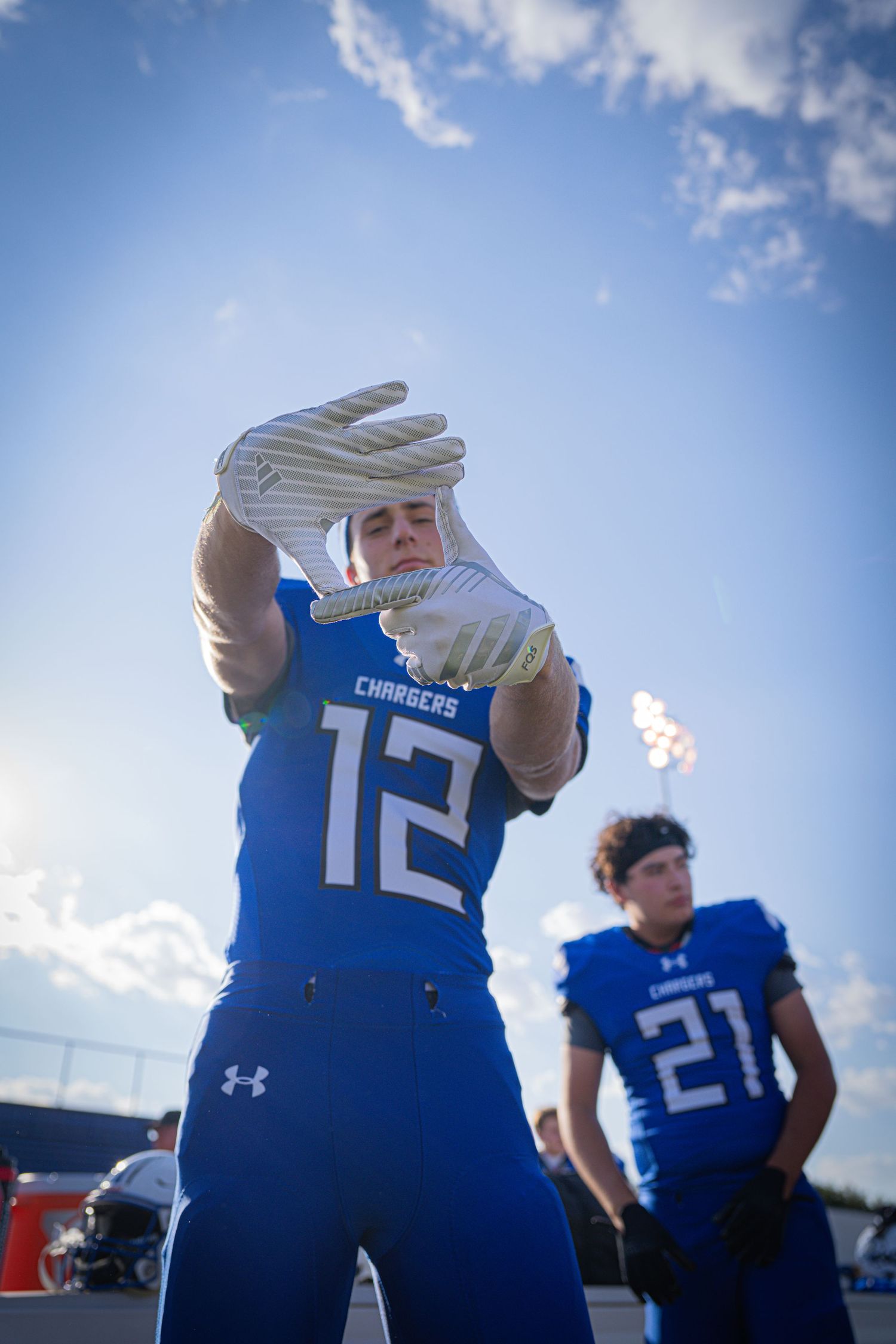 Football player #12 in blue Chargers uniform poses with gloved hands raised on sunny field.