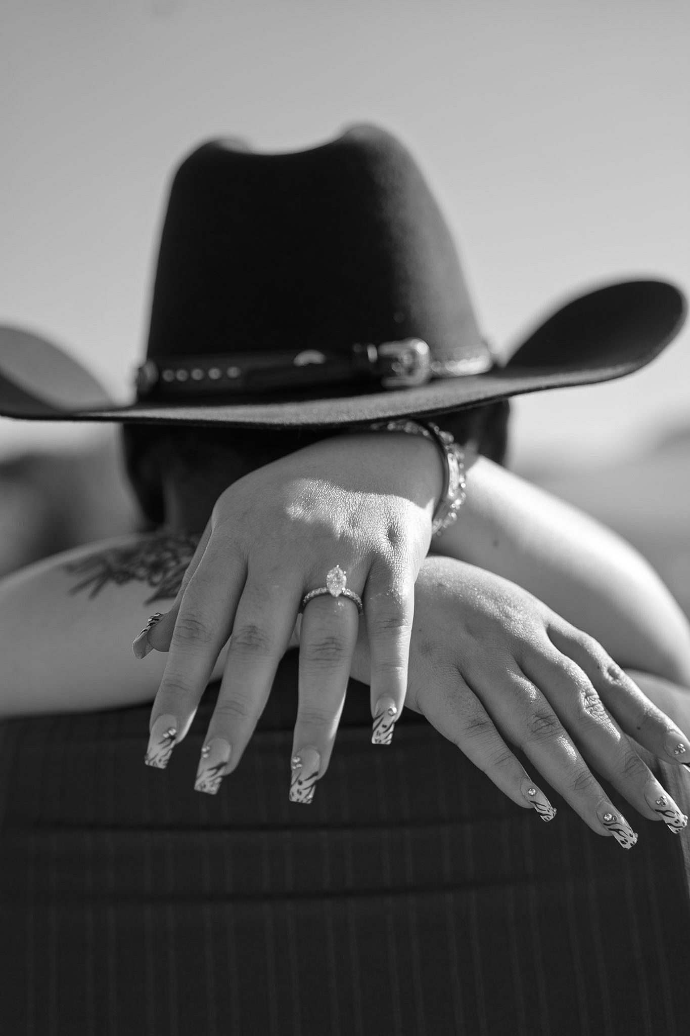 Black and white photo of hands with engagement ring resting on mans shoulder wearing cowboy hat.