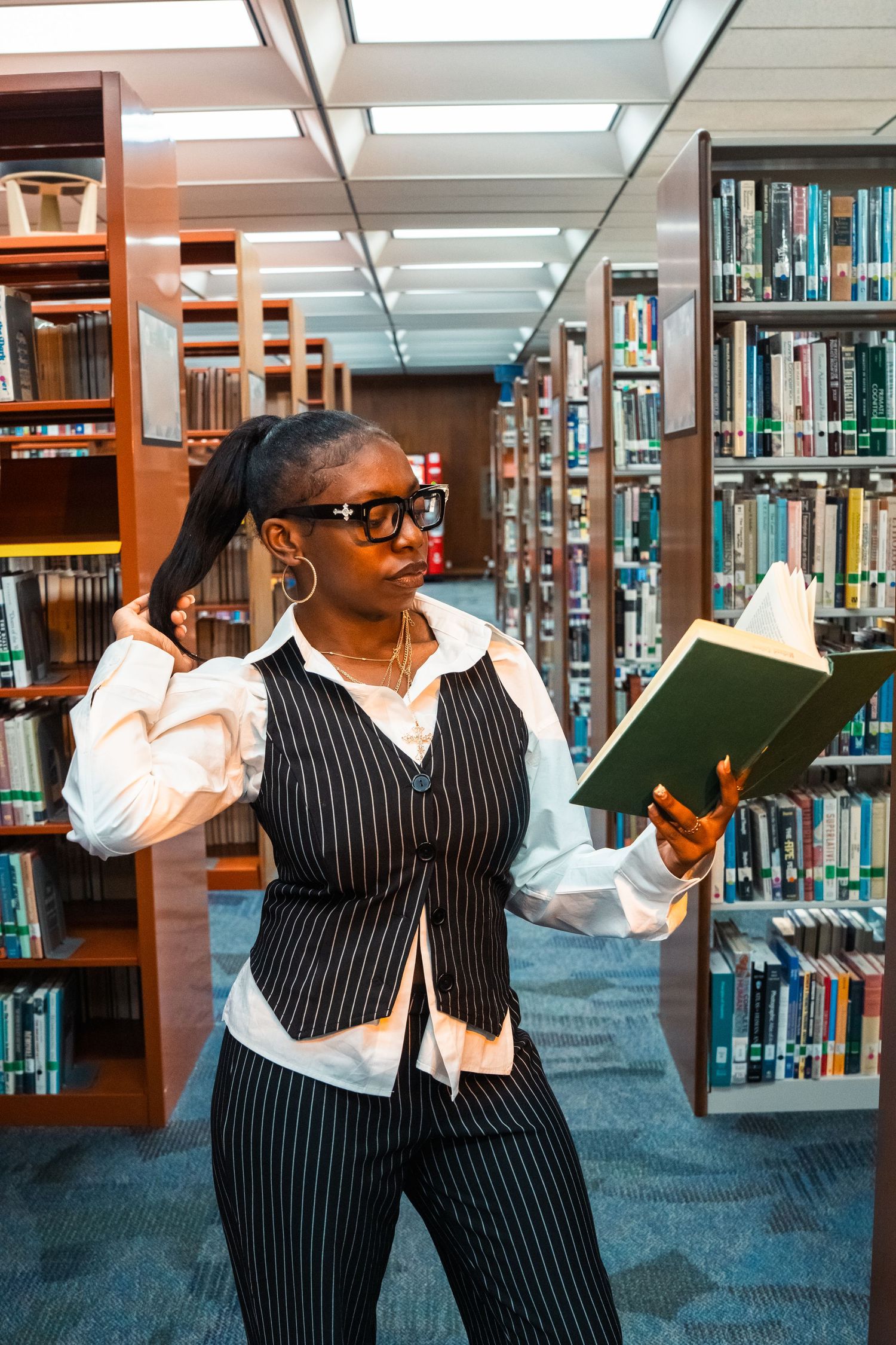 Stylish woman in pinstripe vest reads book while posing dramatically between library bookshelves.