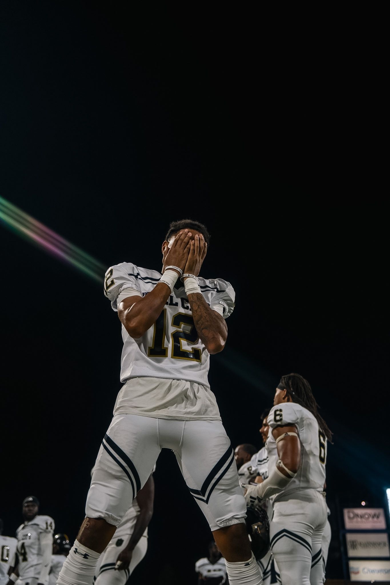 Football player #12 covers face with hands while teammate #6 stands nearby at night game.