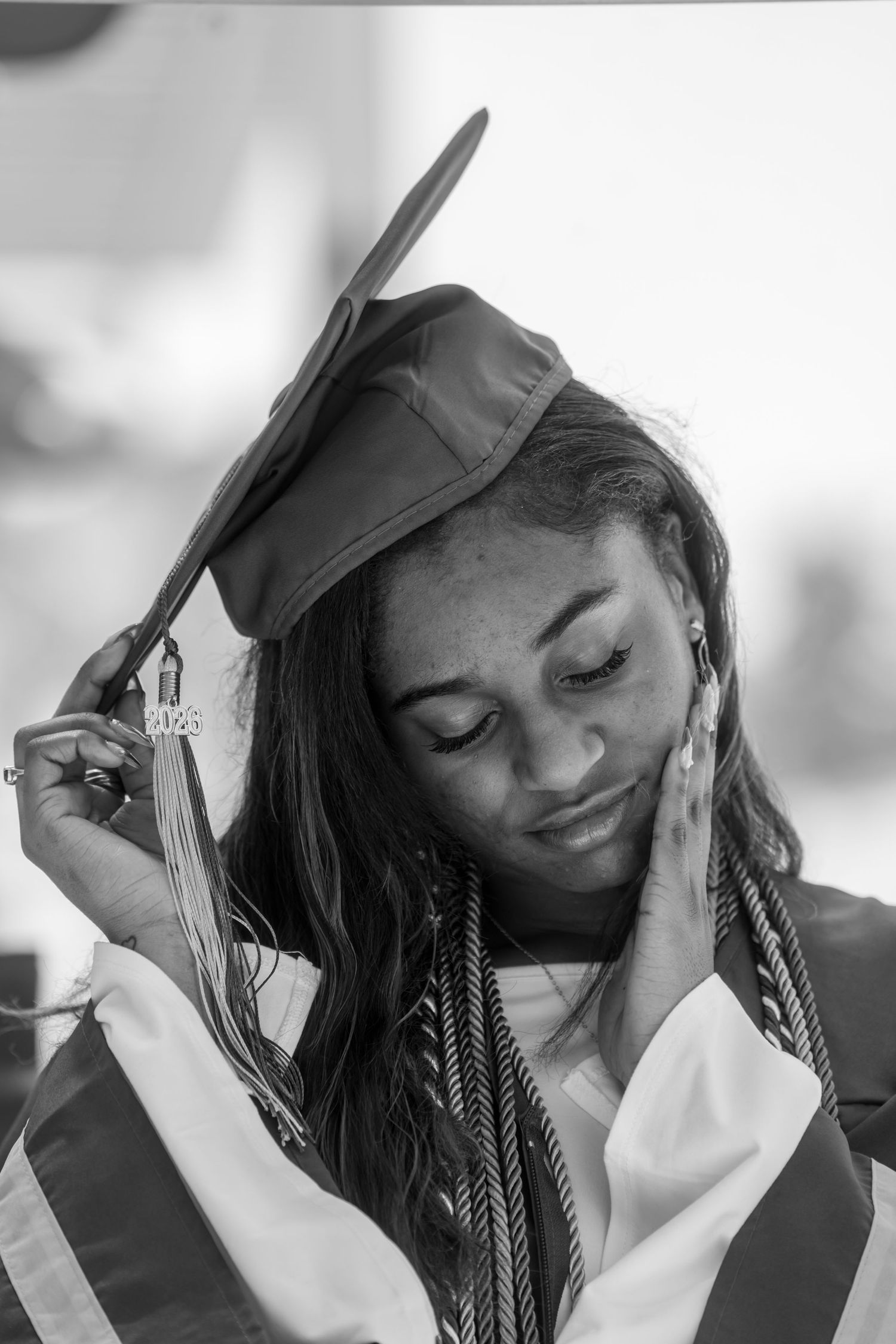 Black and white photo of a young woman in graduation cap and gown, adjusting her mortarboard with honor cords.