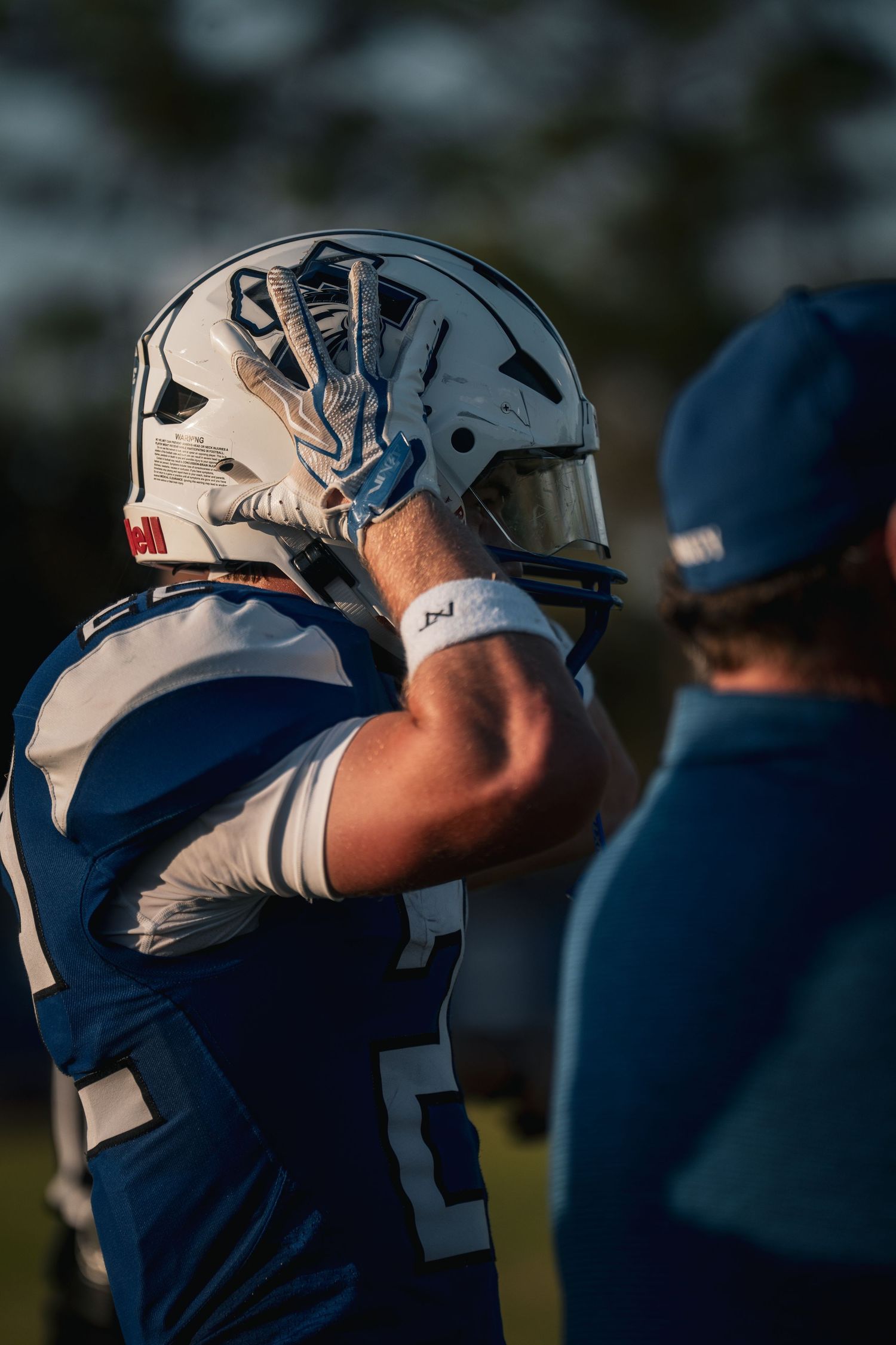A football player in a blue and white uniform adjusts his helmet on the sideline during a game.