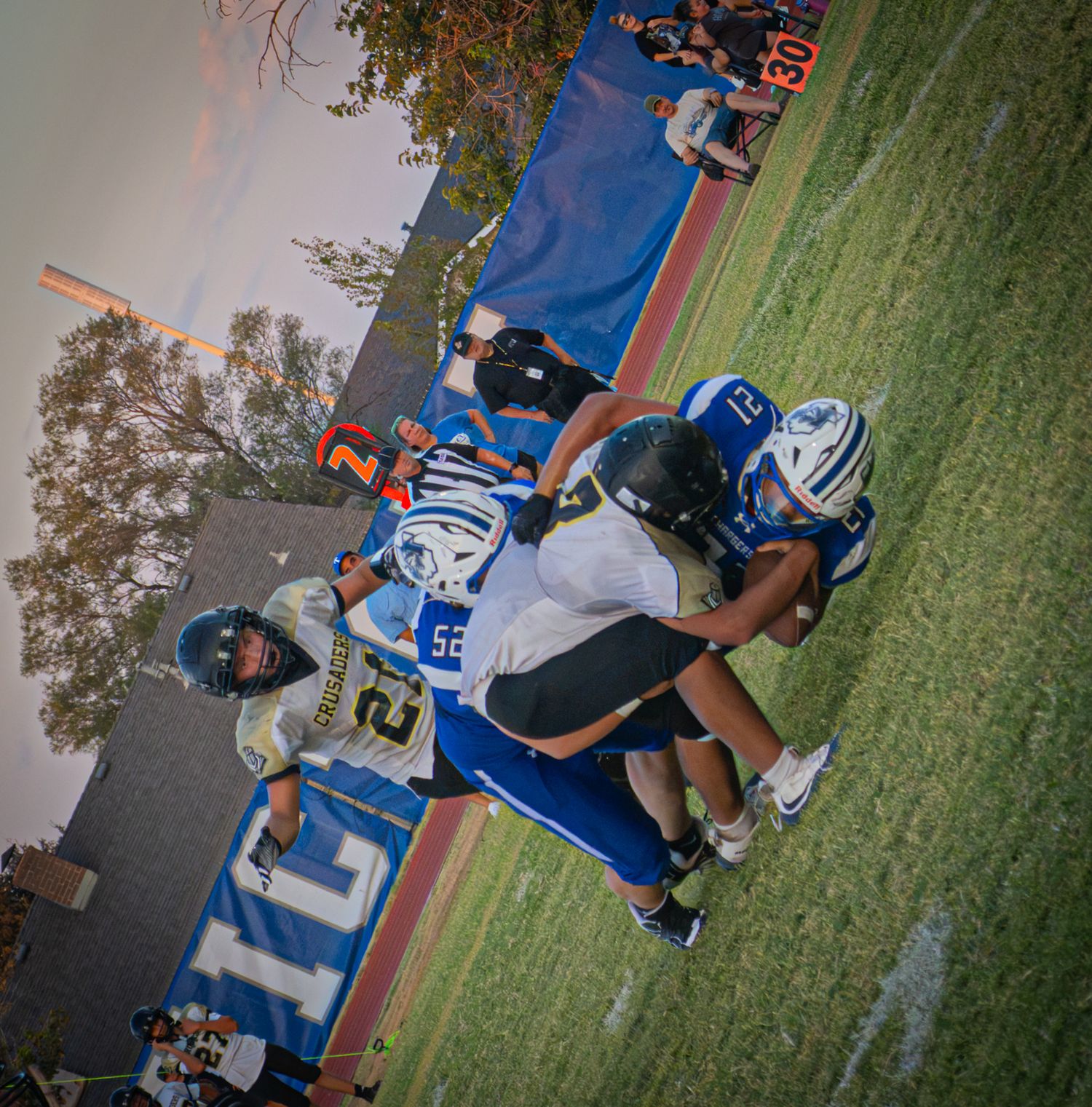 High school football players in blue and white uniforms compete during a game on a red track field surrounded by spectators.