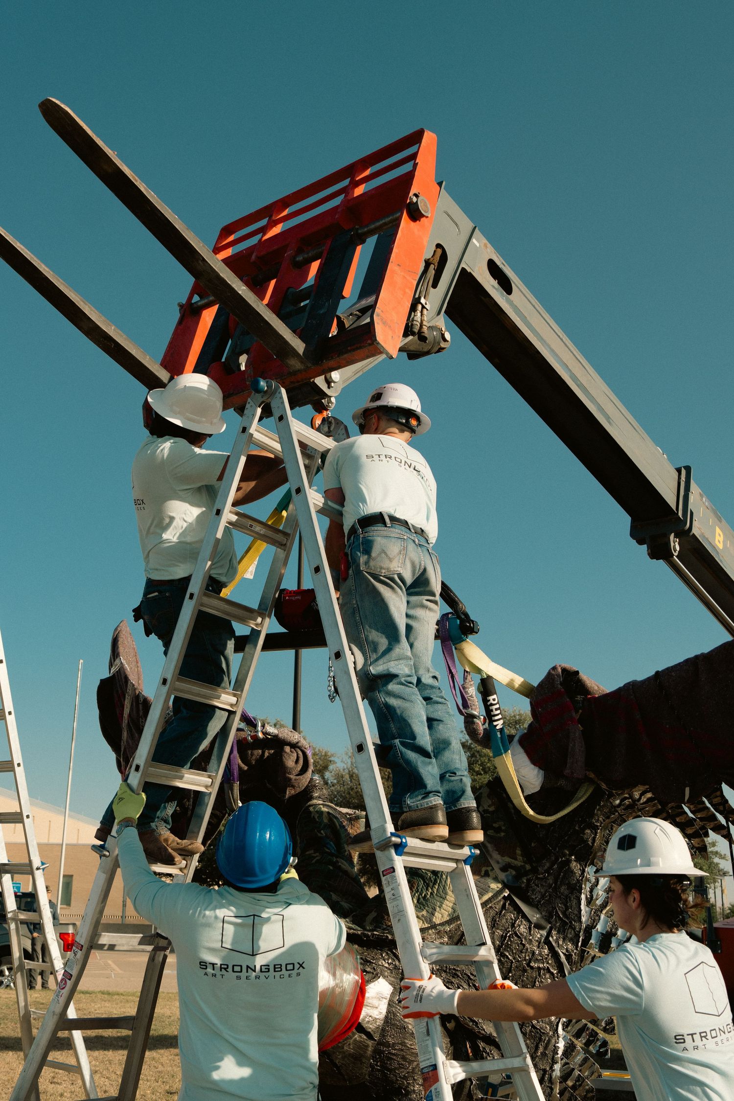 Construction workers in hard hats assembling a large metal sculpture using ladders and a lift.
