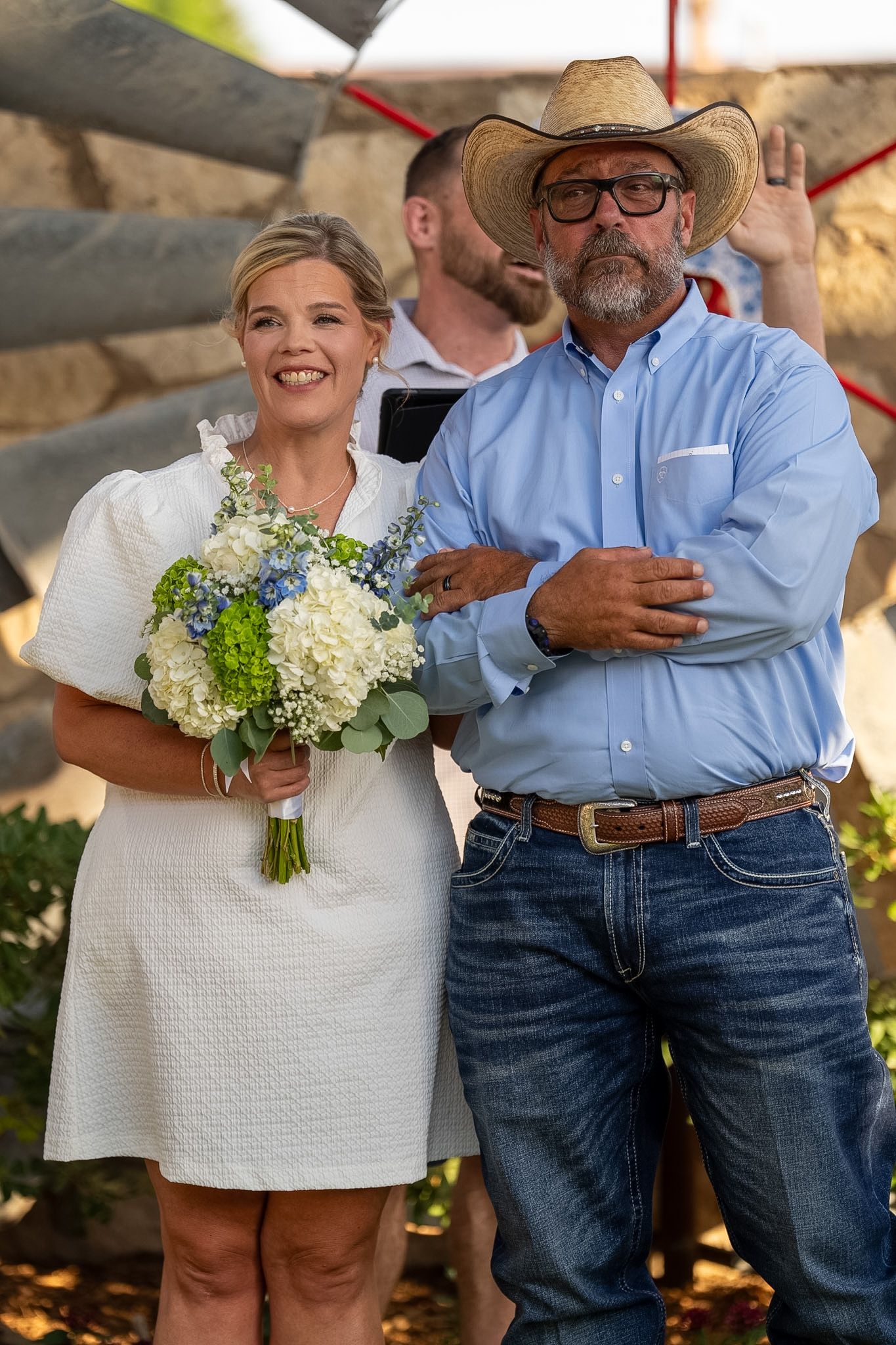 Bride in white dress holding bouquet of white and blue flowers stands with man in cowboy hat at outdoor wedding.