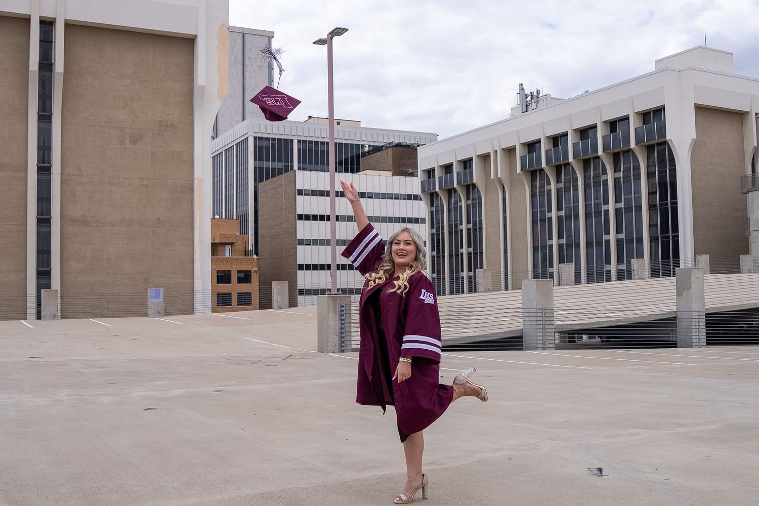 A smiling graduate in maroon cap and gown tosses her graduation cap in the air outdoors on a university campus.