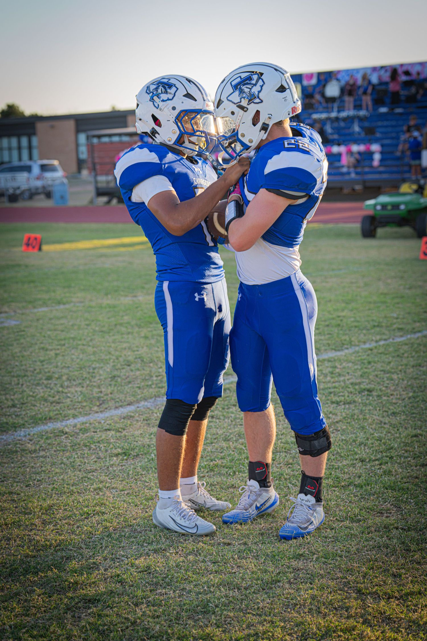 Two blue uniformed football players touch helmets together on field before high school game.