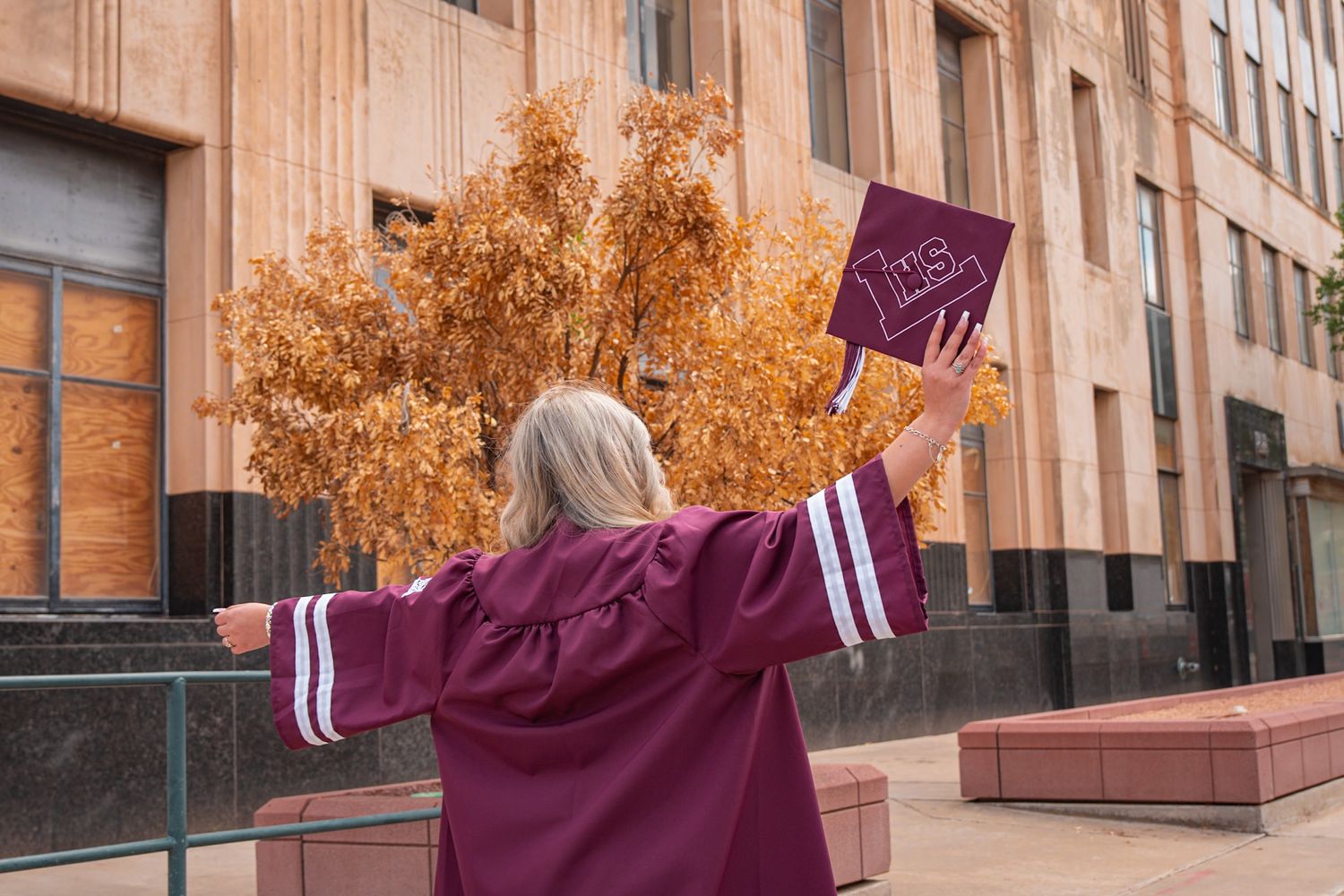 Older woman in maroon graduation gown holding cap with arms outstretched celebrating outdoors near building with autumn trees.