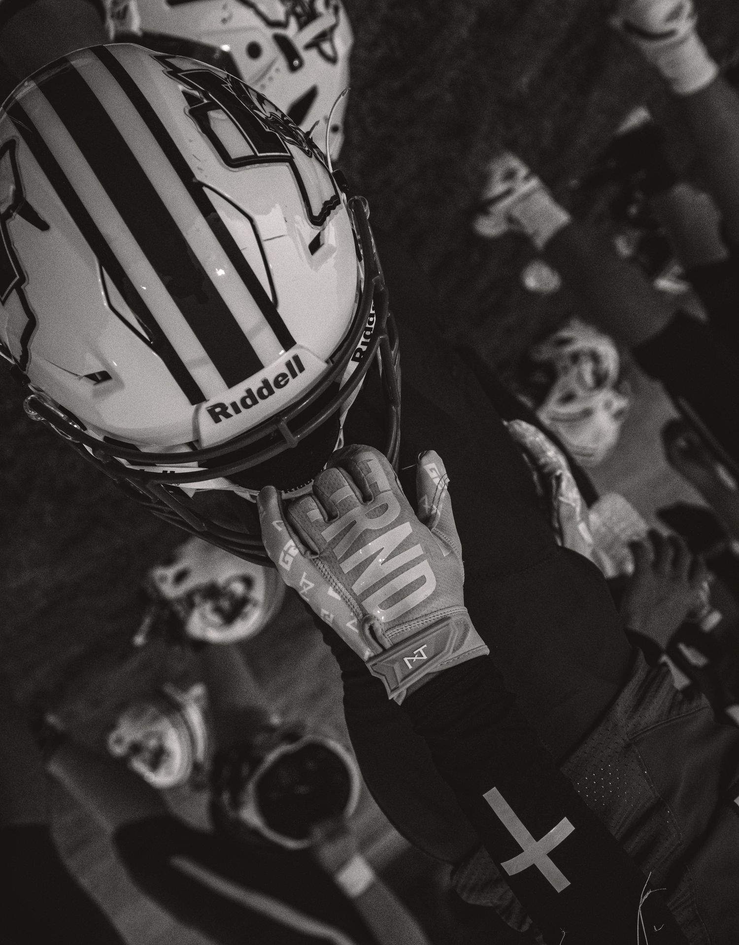 Black and white photo of a football player holding a Riddell helmet while wearing gloves in a locker room.