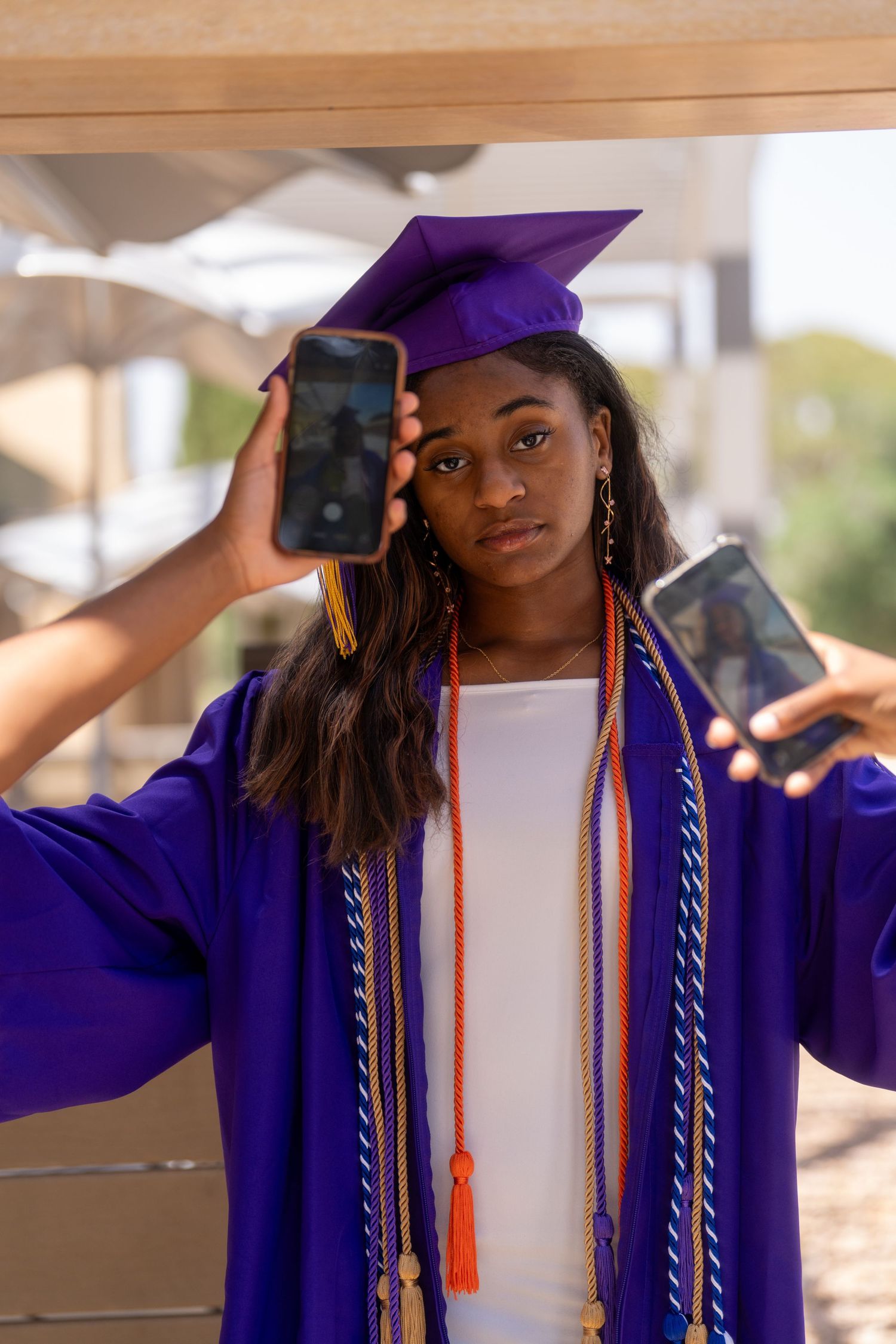 Young Black woman in purple graduation cap and gown holds two smartphones up beside her head outdoors.