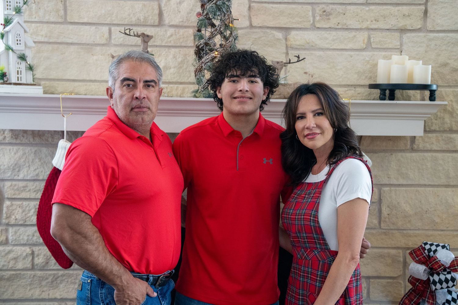 Family of three posing in red outfits by a decorated fireplace during Christmas holiday celebrations.