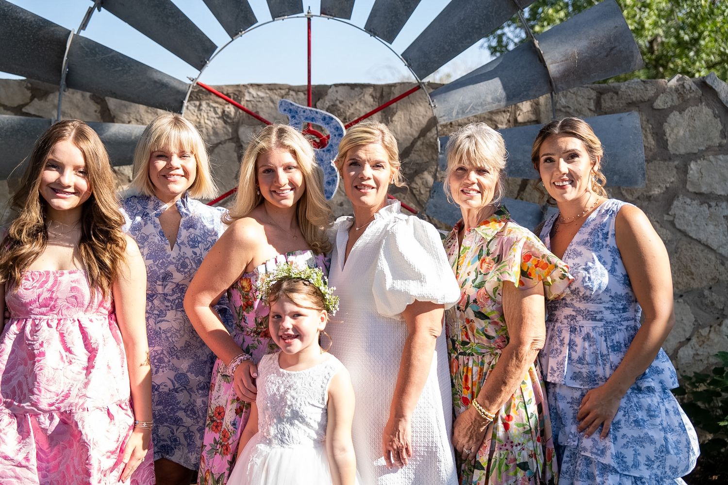 Bride in white dress poses with bridesmaids in floral dresses and flower girl outdoors near stone wall.