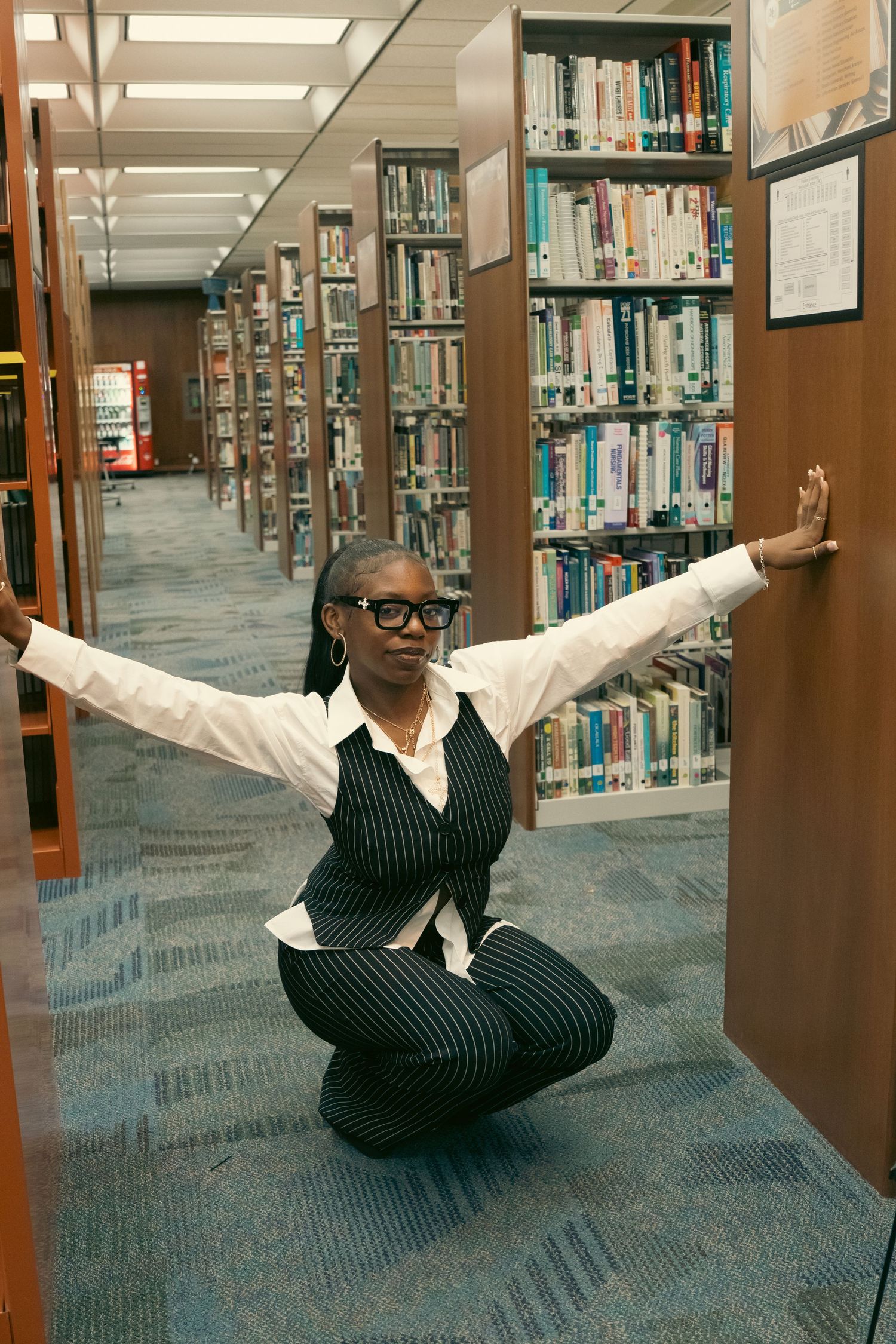 Black woman in pinstripe suit crouches between library shelves with arms outstretched in a dynamic pose.