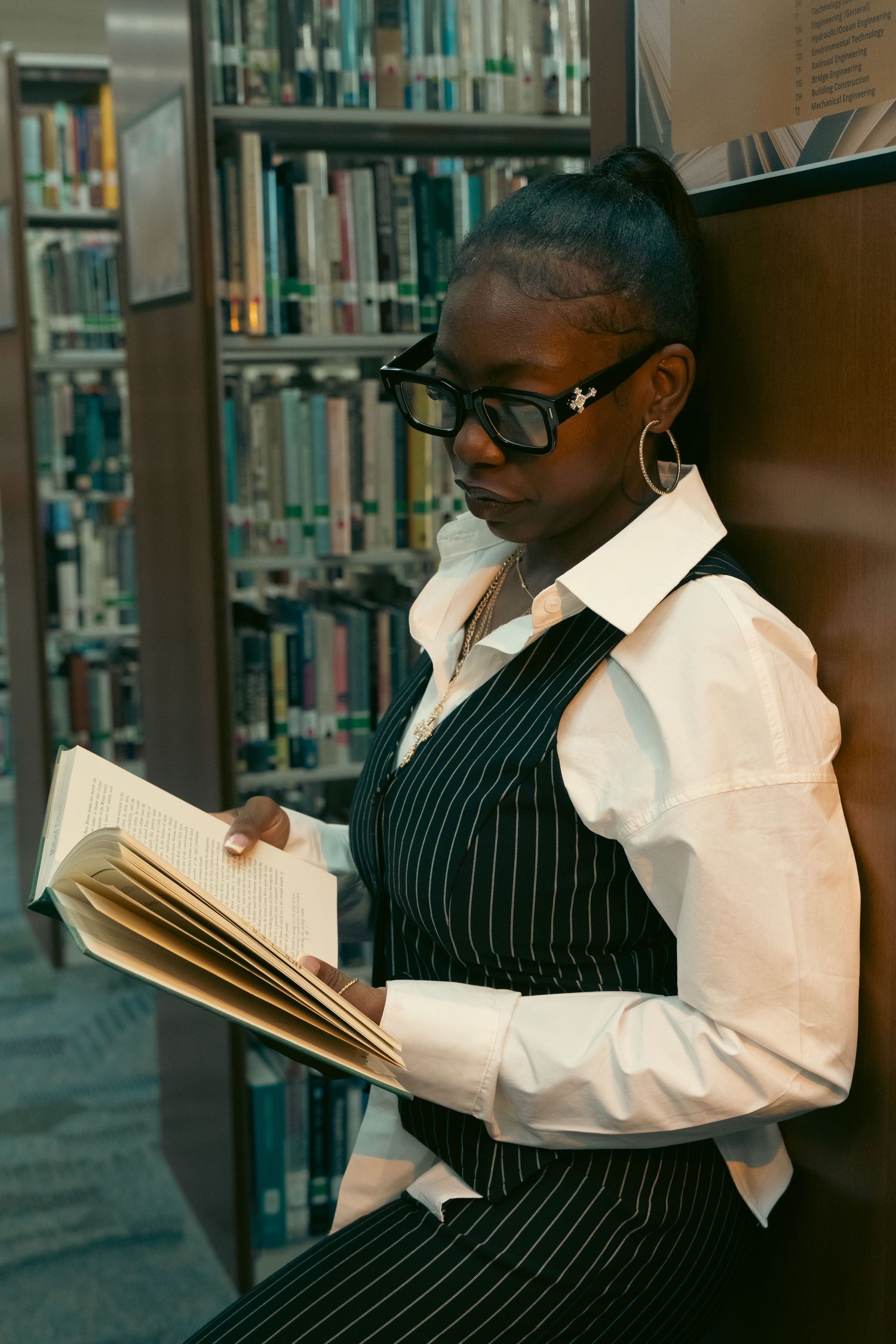 Young Black woman in pinstripe vest reads book leaning against library shelf.