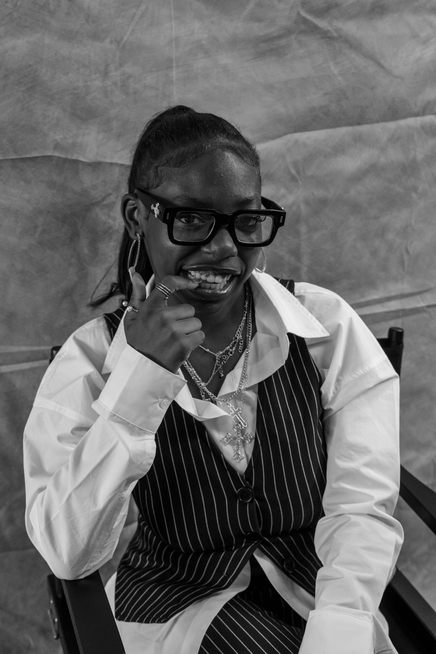 Smiling young Black woman wearing glasses and pinstripe vest over white shirt, posing in black and white portrait photo.
