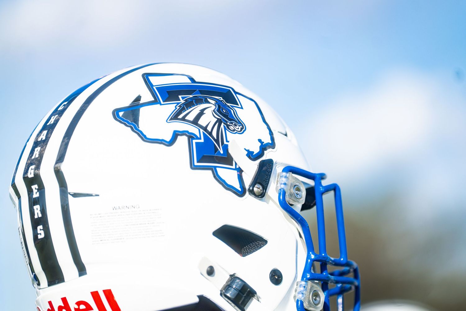 White Riddell football helmet with blue Trojans logo and blue face mask against a sky background.