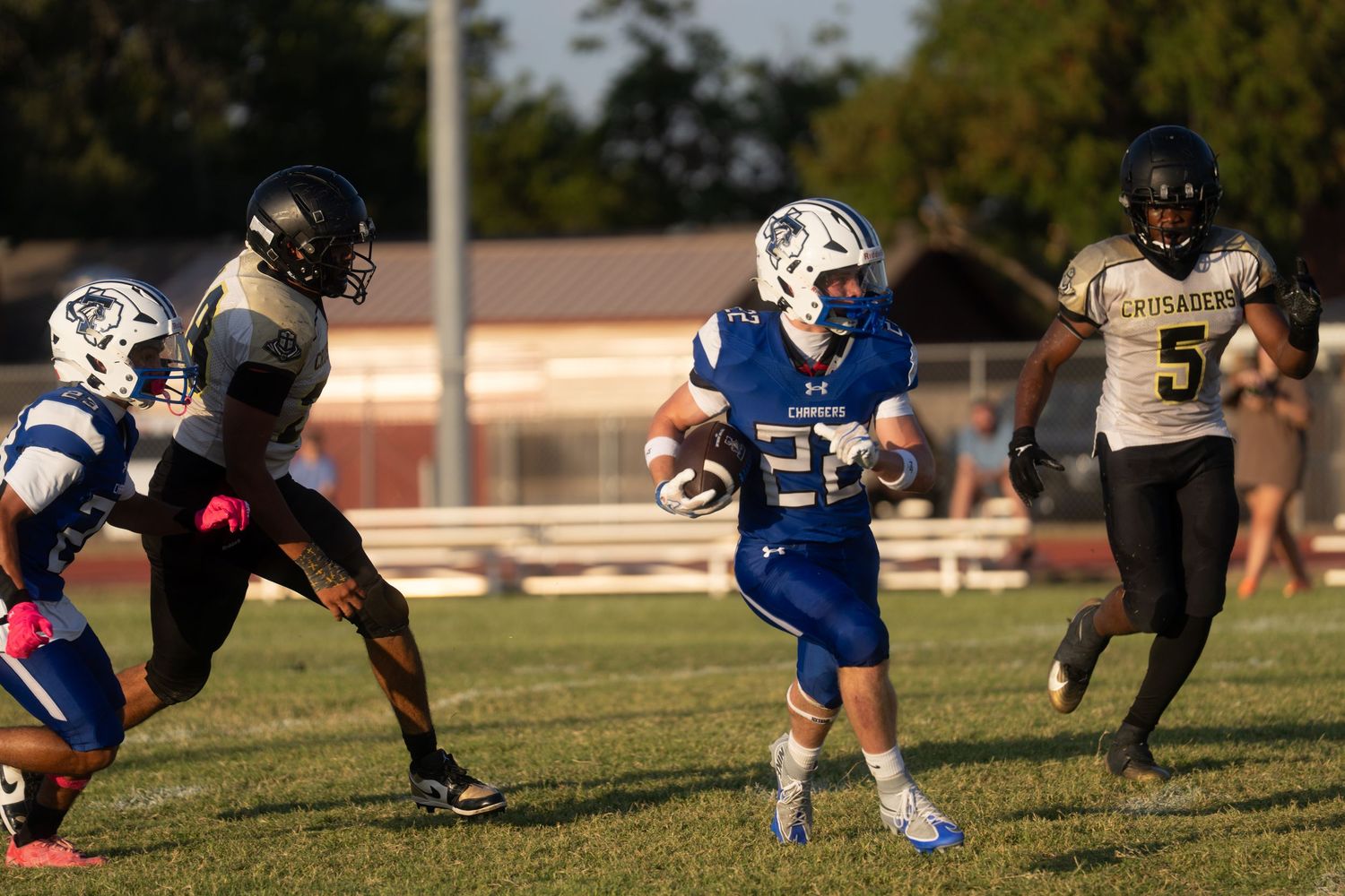 High school football player in blue uniform runs with ball during evening game against black-uniformed opponents.