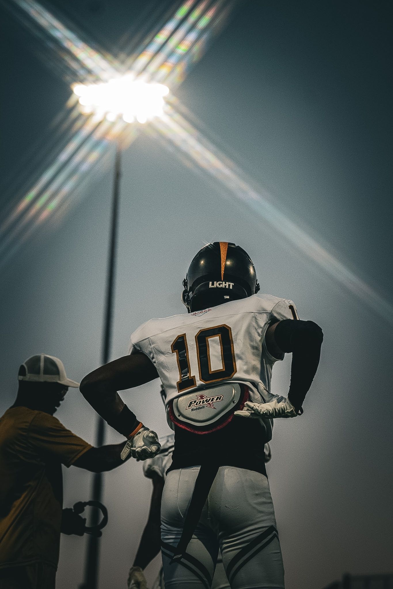 Football player number 10 in a Light helmet stands under bright stadium lights holding a football.