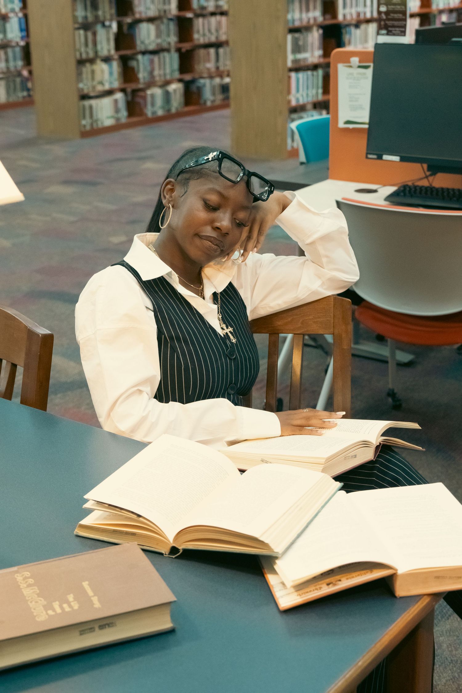 Black woman in pinstripe vest rests head on hand while studying open books at a library table.