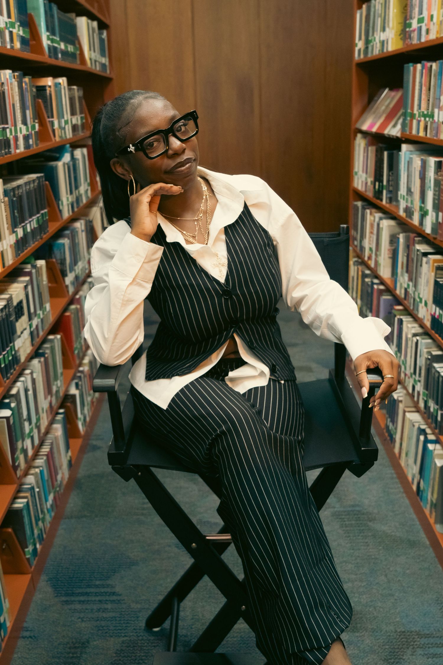Stylish Black woman in pinstripe suit poses thoughtfully in director chair between library shelves.