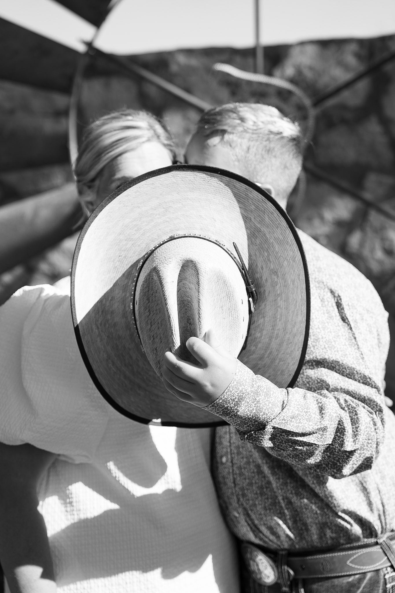Black and white photo of a couple embracing, with a child holding a cowboy hat between them near a windmill.
