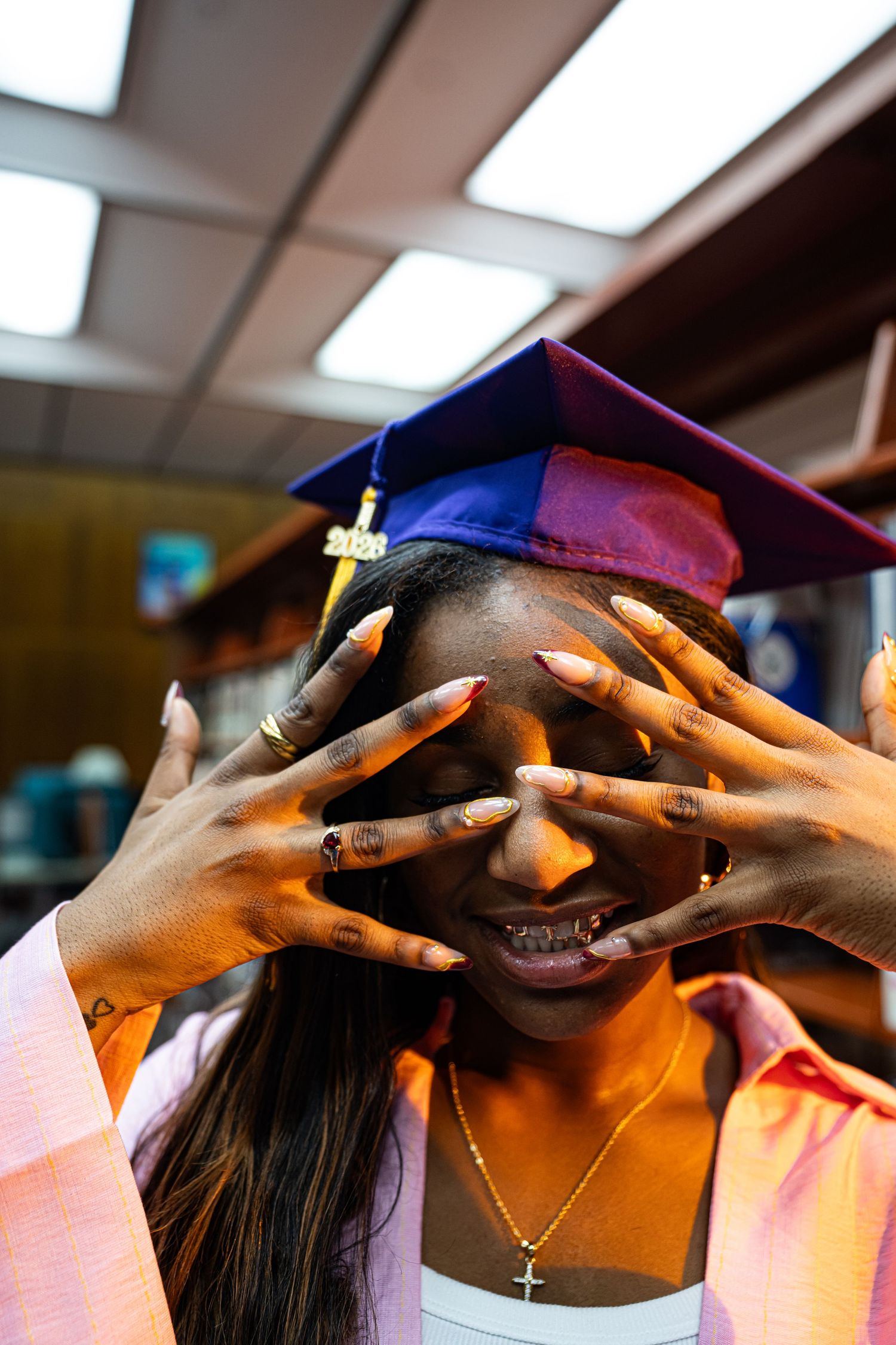 Graduate in blue cap and gown poses playfully covering face with decorated nails, wearing cross necklace.