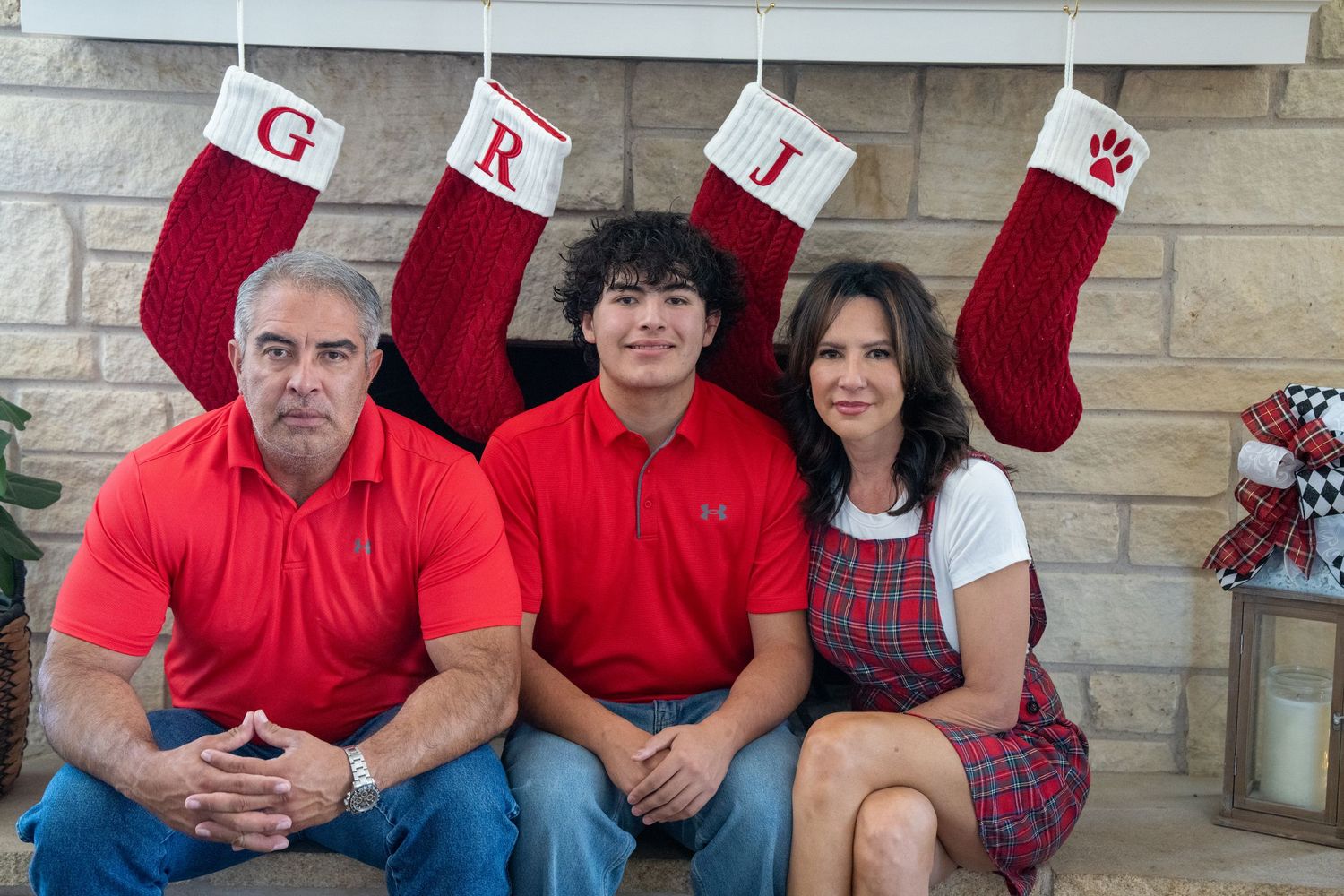 Family of three posing in front of a fireplace with Christmas stockings labeled G, R, and J during the holiday season.