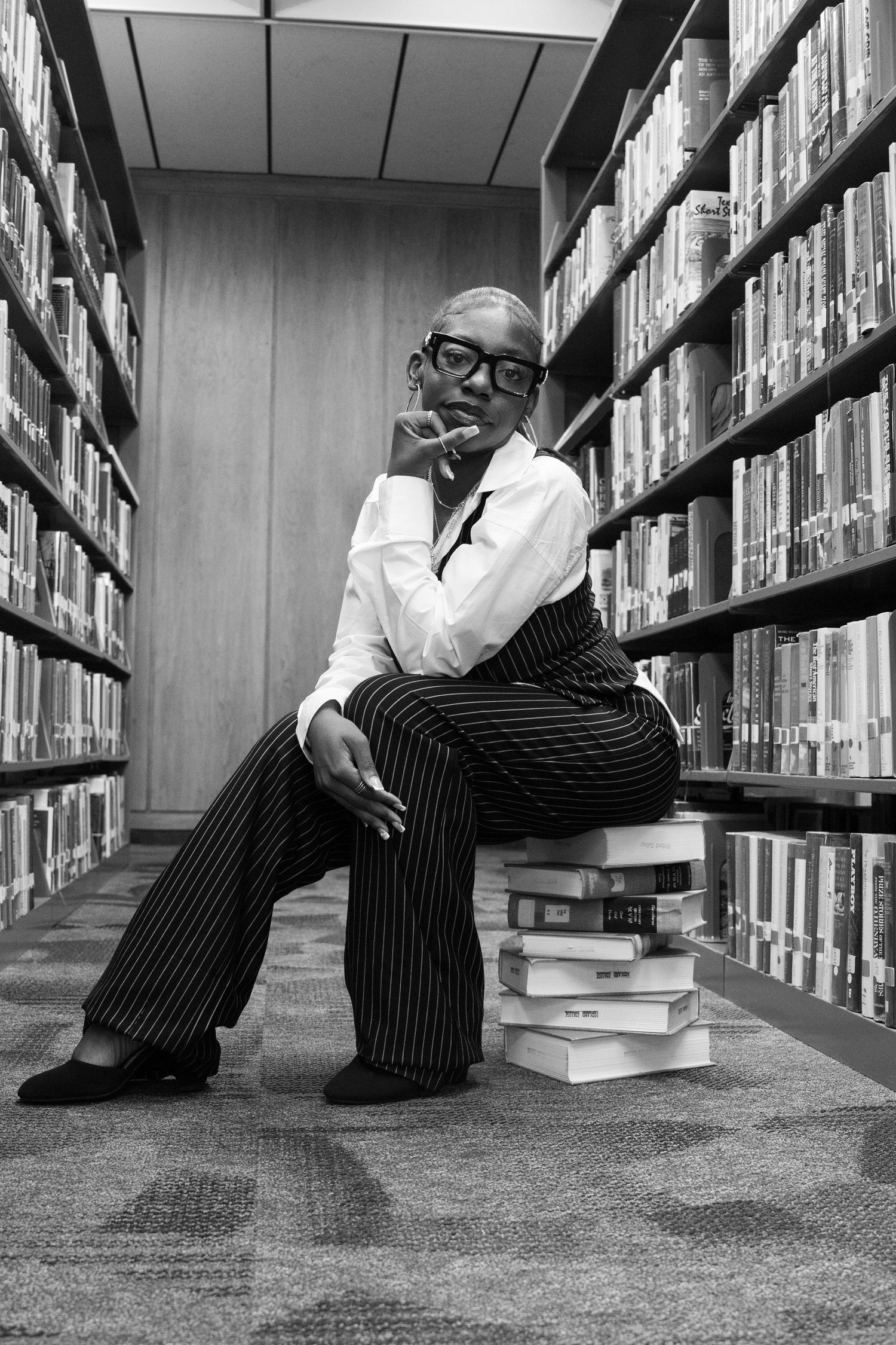 Black and white portrait of elegant woman in pinstripe trousers sitting on stacked books in library aisle.