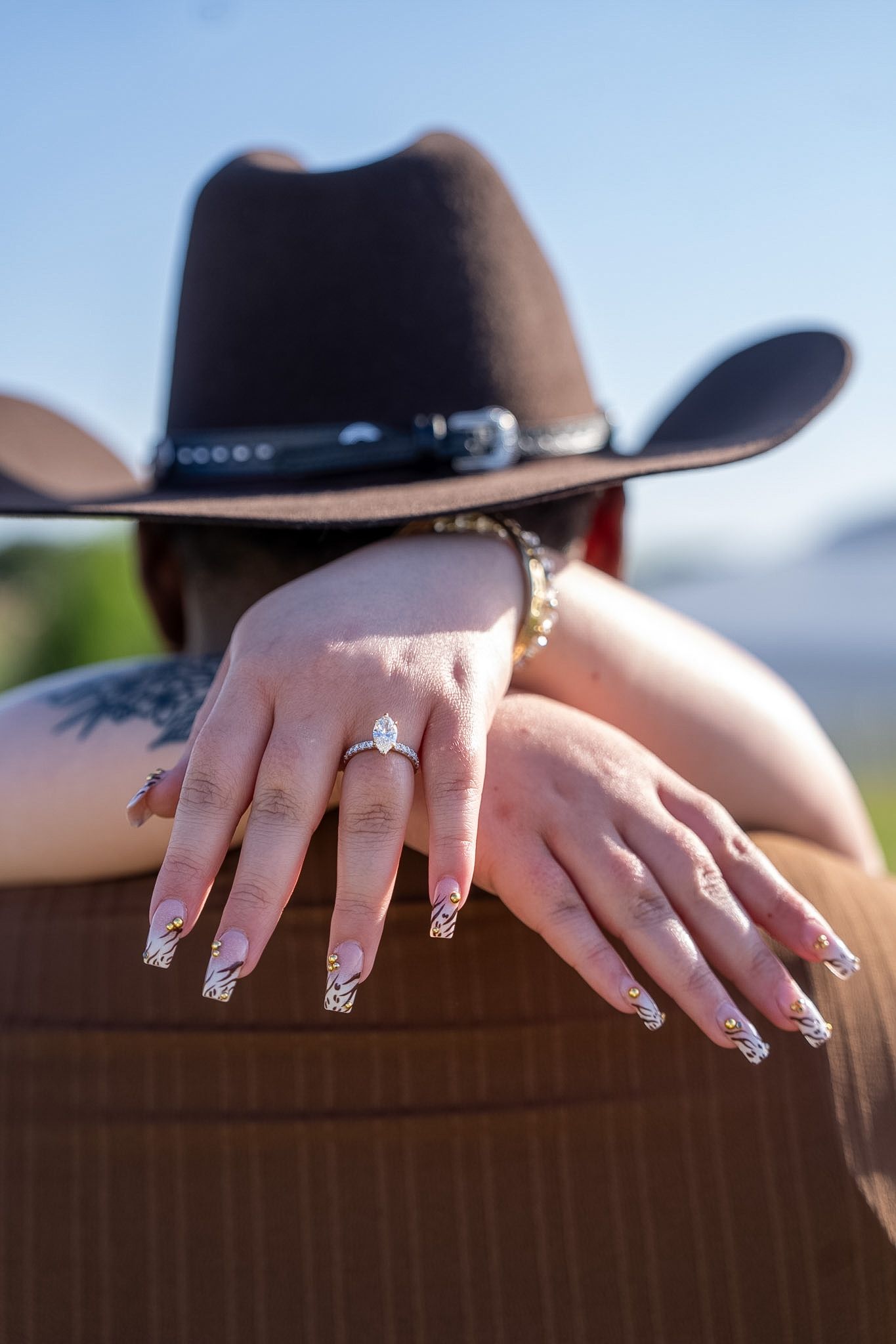 Close-up of womans hands with marquise engagement ring resting on mans shoulder in cowboy hat.