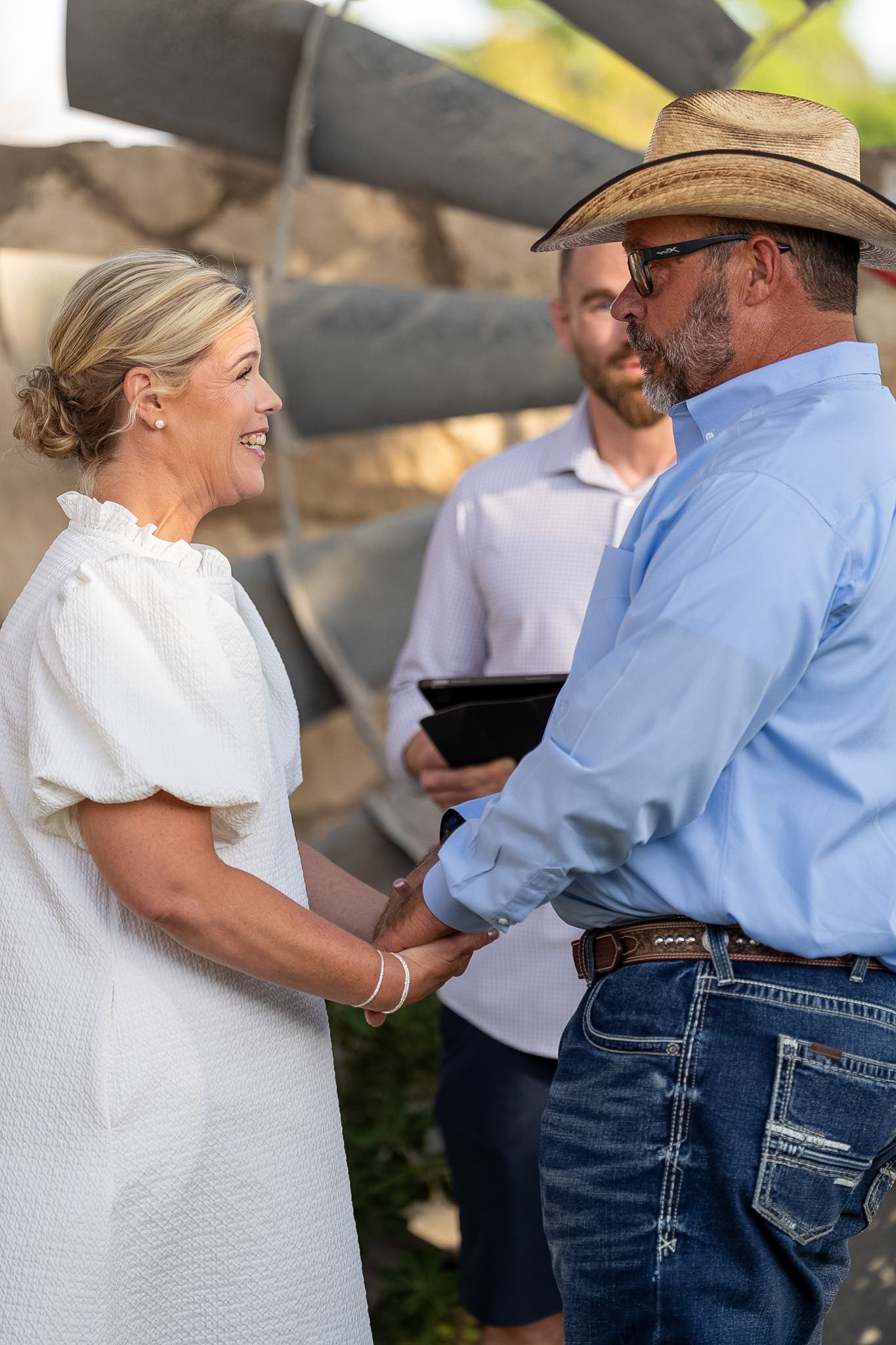 Couple holds hands during outdoor wedding ceremony while officiant reads vows behind them.