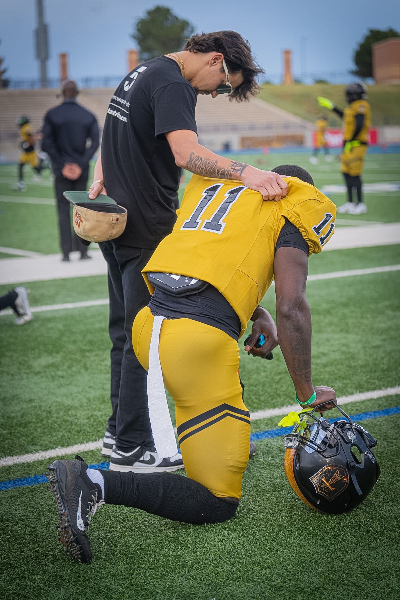 Athletic trainer assists kneeling football player #11 in gold jersey on green turf field.