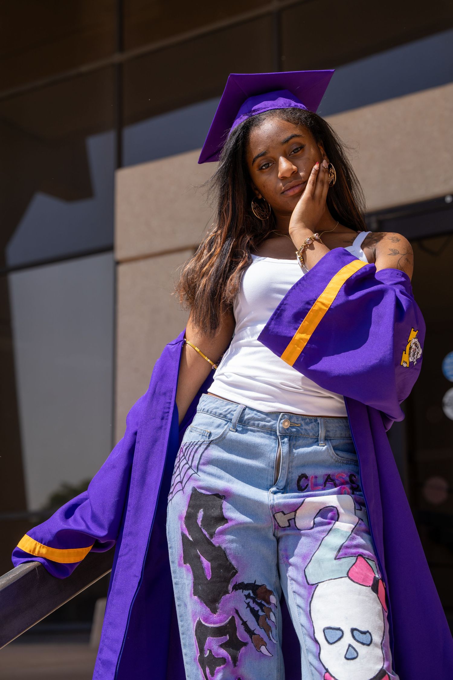 Young Black woman in purple graduation cap and gown with custom painted jeans posing outdoors.