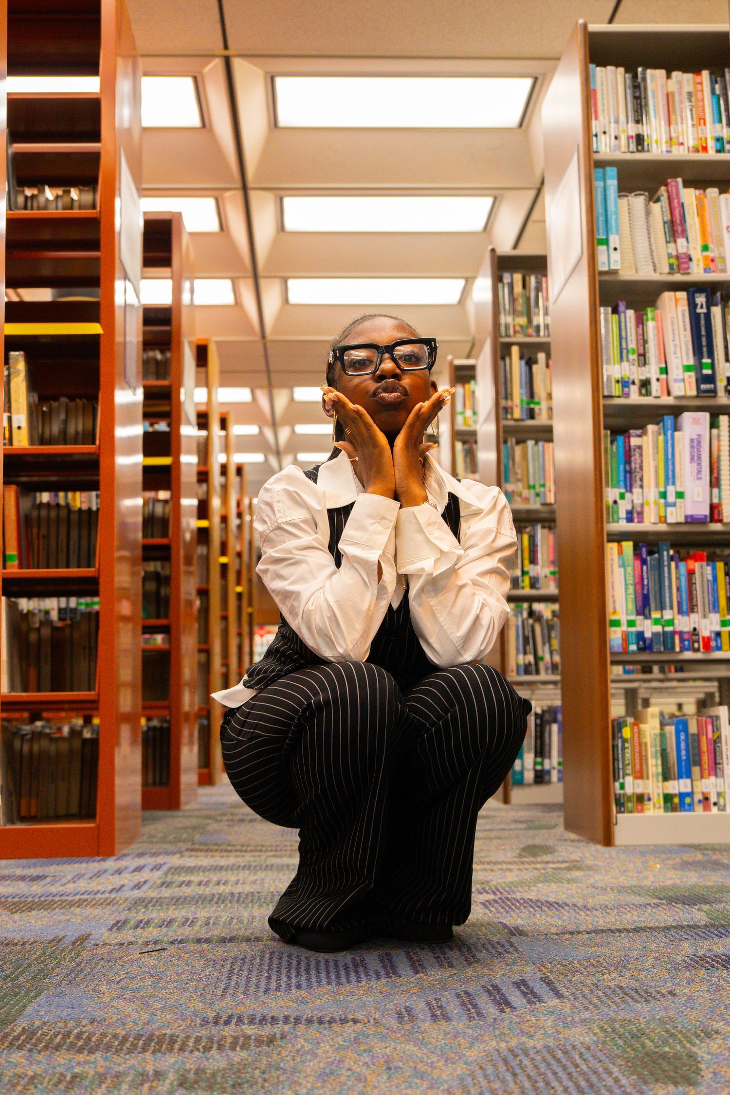 A young Black woman in glasses and striped overalls poses thoughtfully between colorful library bookshelves.