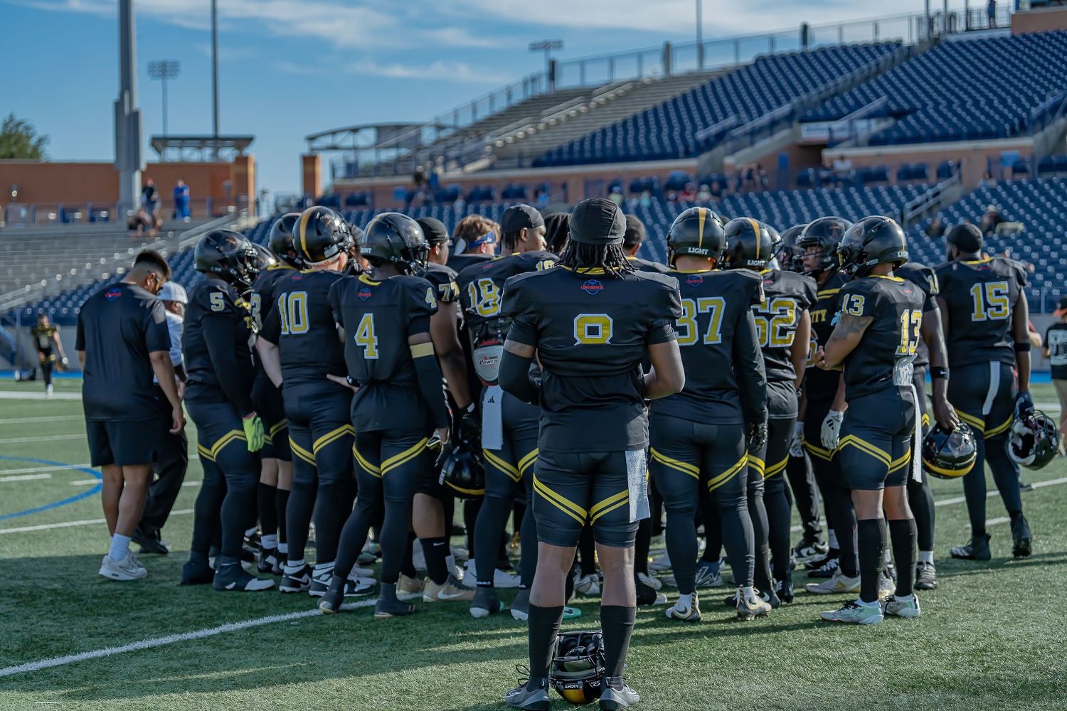 Football team huddles together in black and gold uniforms on stadium field before game.