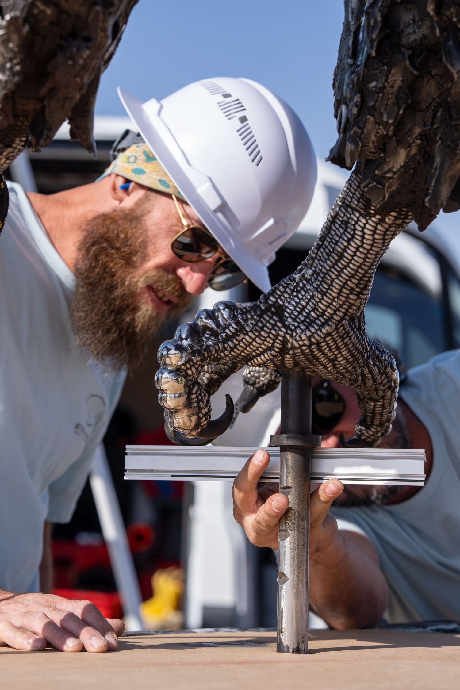 Bearded man in white hard hat carefully assembles a metallic dragon claw sculpture outdoors.