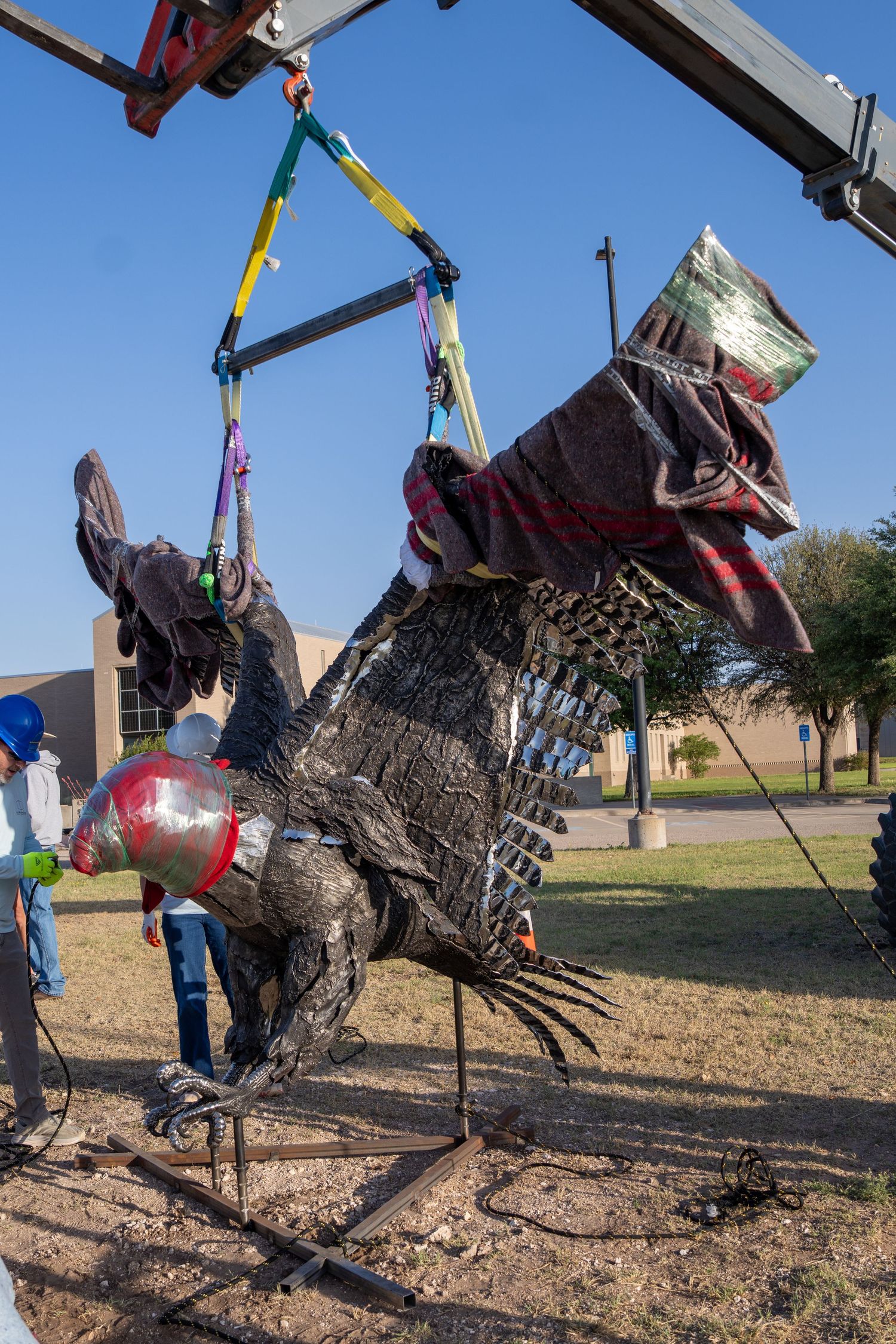 Large metal turkey sculpture being lifted by crane with colorful straps outdoors on a sunny day.