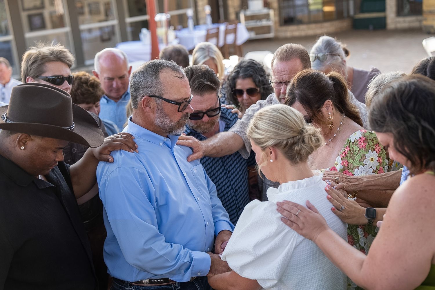 Group of people gathered in prayer circle outdoors, placing hands on couple in center at public event.